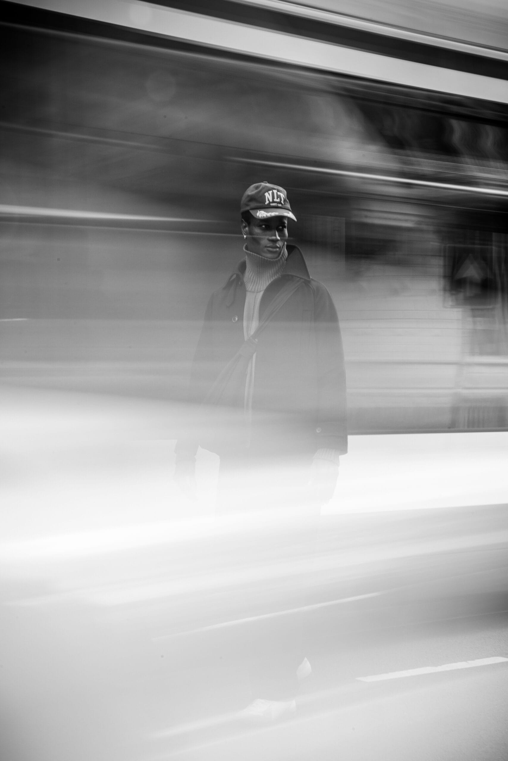 Zwart-wit portret van een man met een pet en coltrui, staand voor een voorbijrazende trein met bewegingsonscherpte