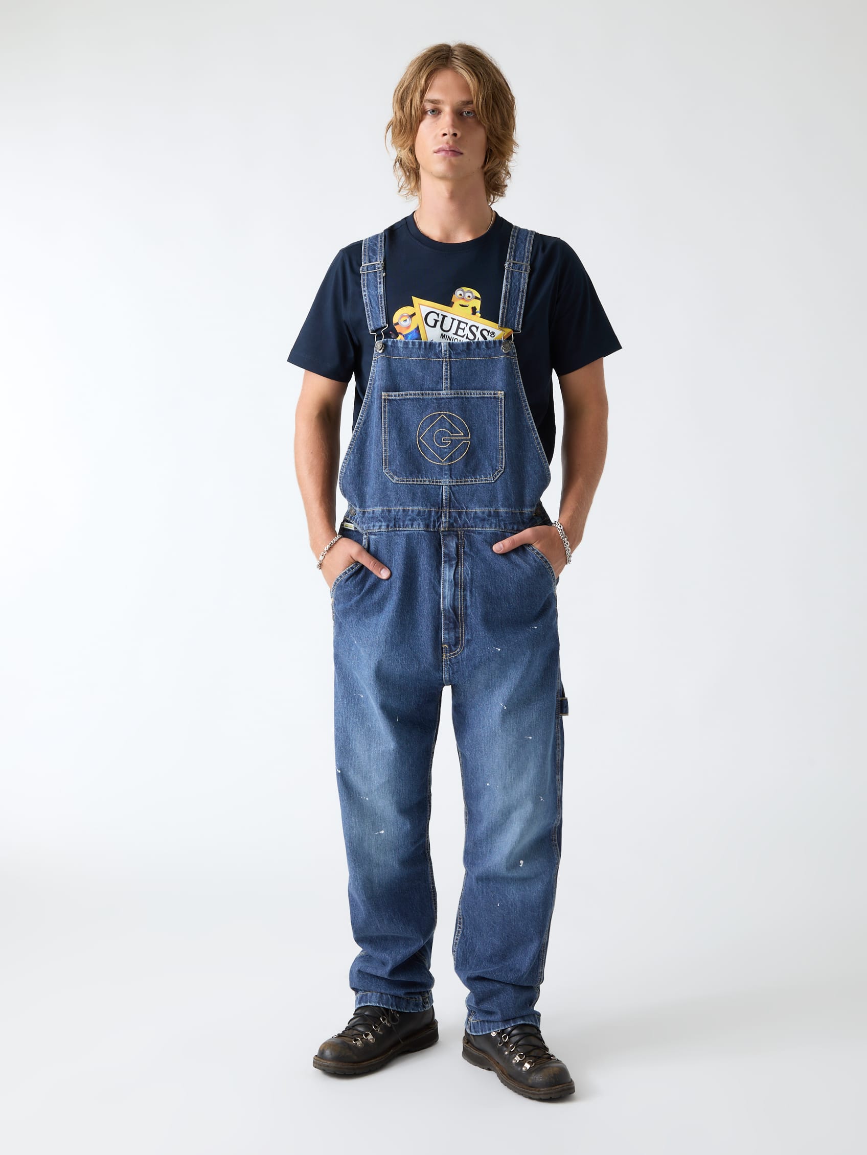 Man met halflang blond haar draagt een blauwe denim tuinbroek van Guess over een donkerblauw T-shirt met een Minions-print