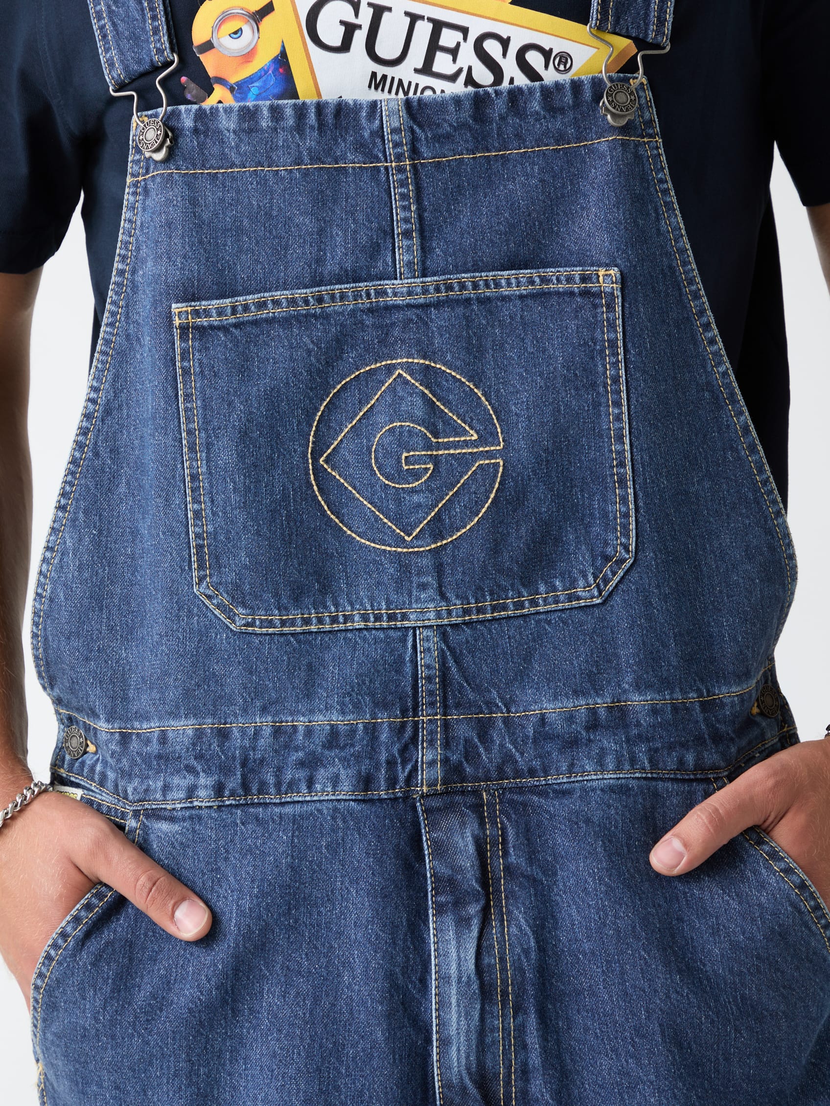 Close-up van een blauwe denim tuinbroek van Guess met een geborduurd Minions-logo op de borstzak