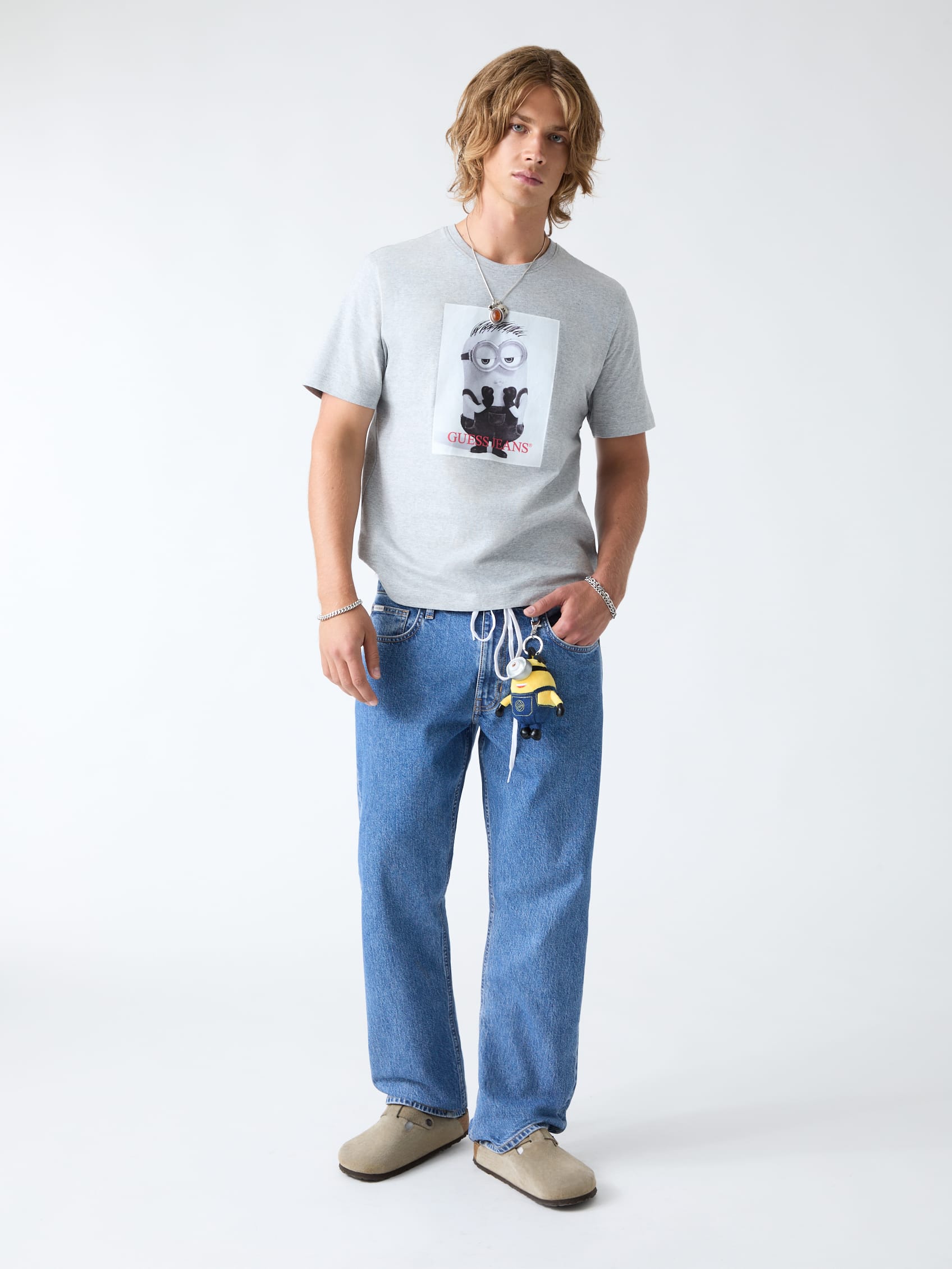 Man in een grijs T-shirt met Minion-print, een blauwe jeans en beige instappers allemaal van het modemerk Guess