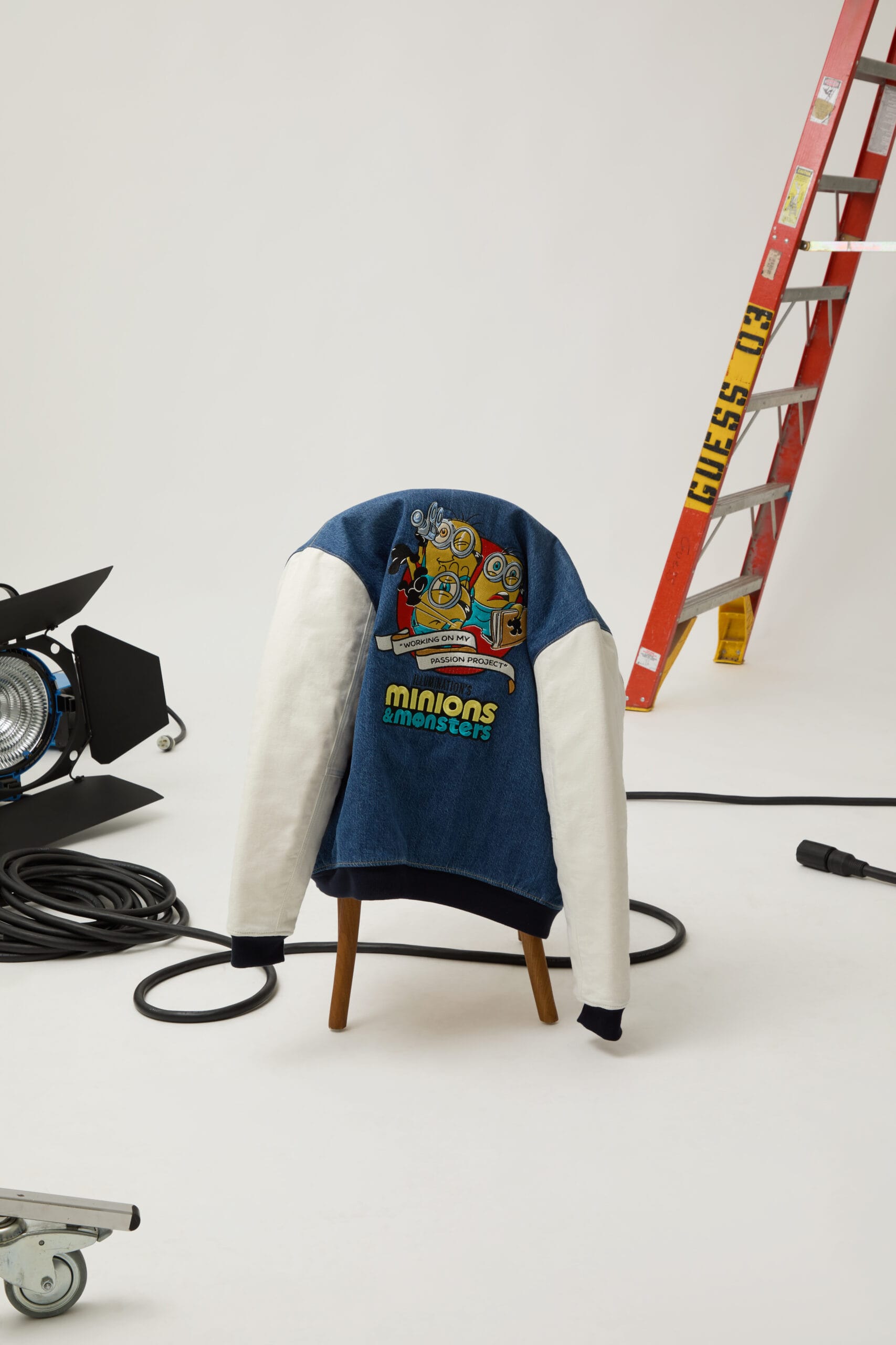 Achterkant van een denim varsity jack van Guess met Minions-illustratie en de tekst Minions & Monsters op een kruk in een studio
