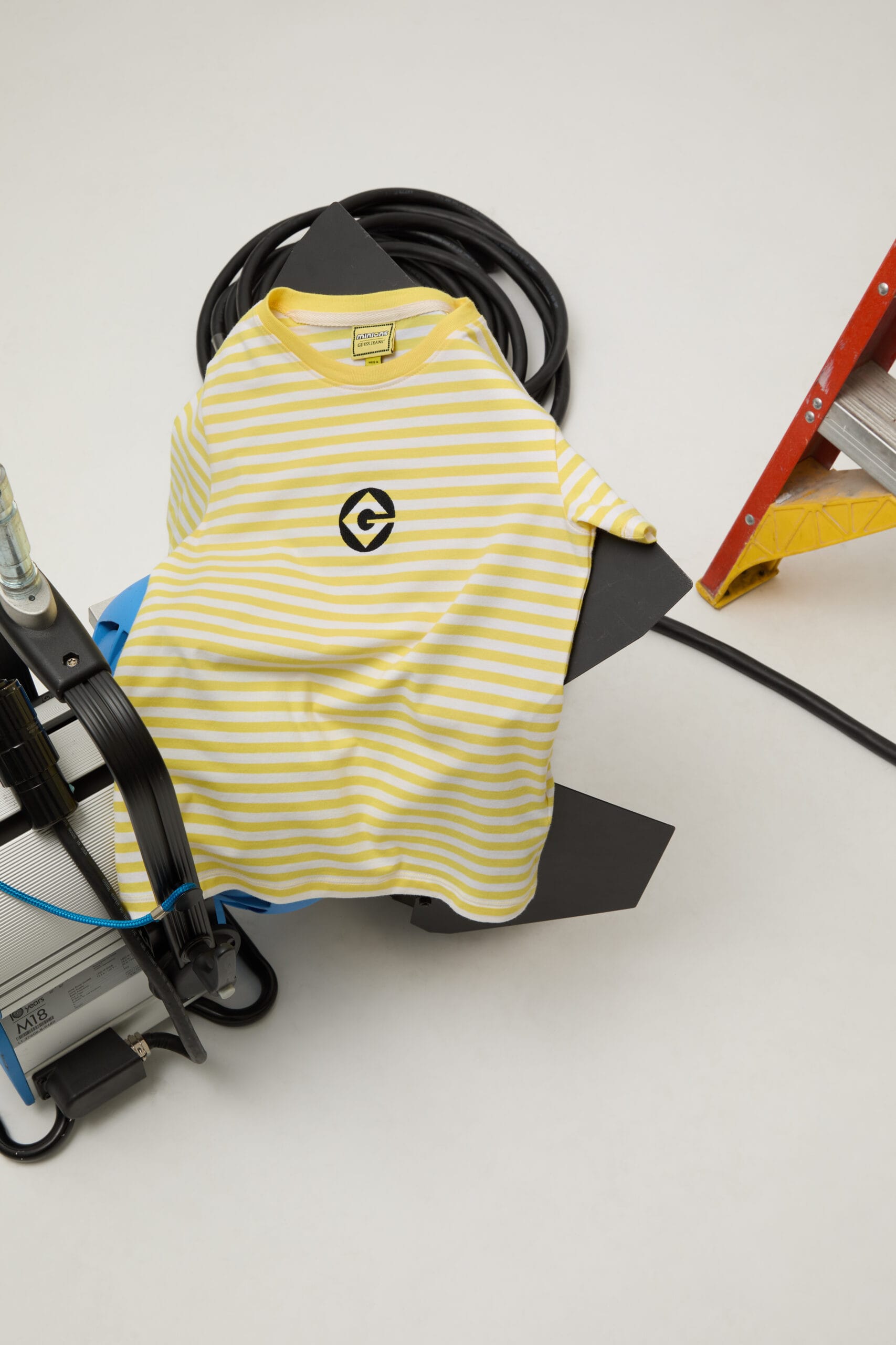 Geel-wit gestreept T-shirt van Guess met een Minion-logo op de borst, gedrapeerd over studio-apparatuur