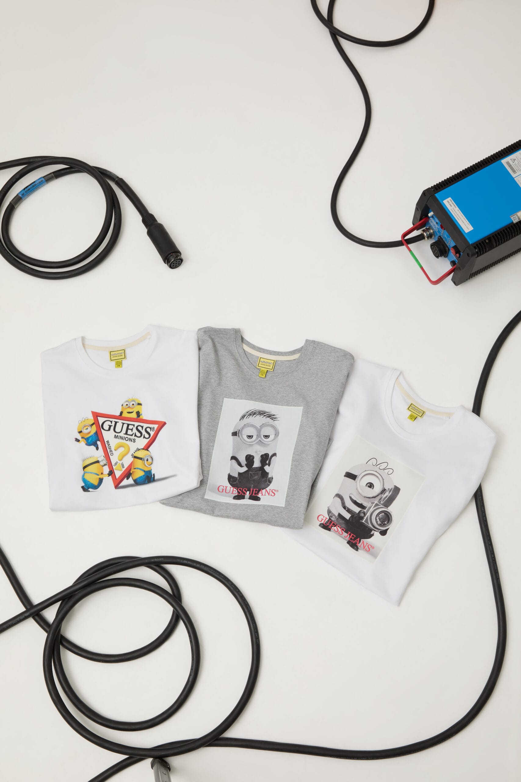 Drie Guess Jeans T-shirts met Minions-prints, omringd door zwarte kabels en technische apparatuur