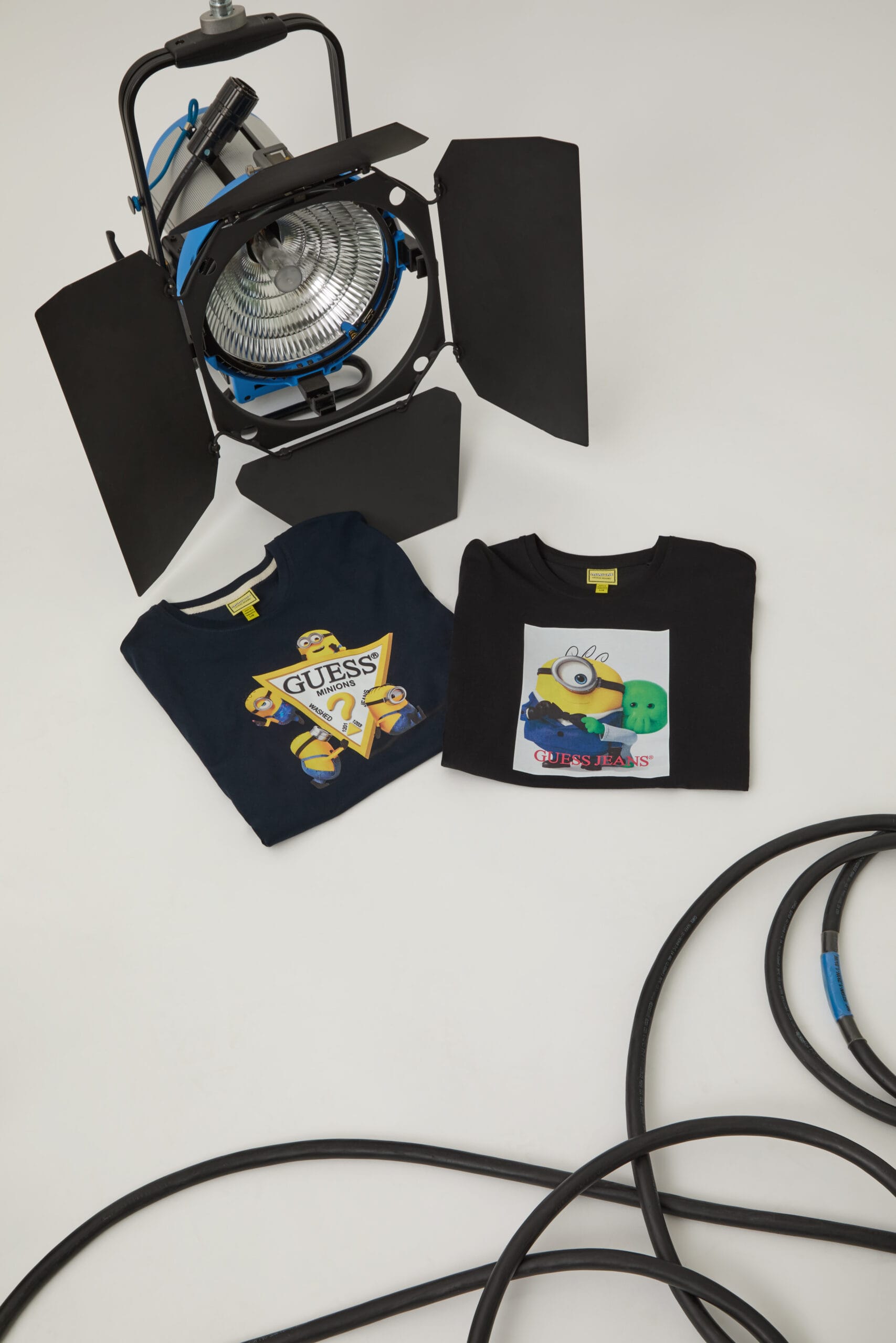 Twee opgevouwen Guess t-shirts met Minions-prints naast een grote studiolamp en zwarte kabels op een witte achtergrond