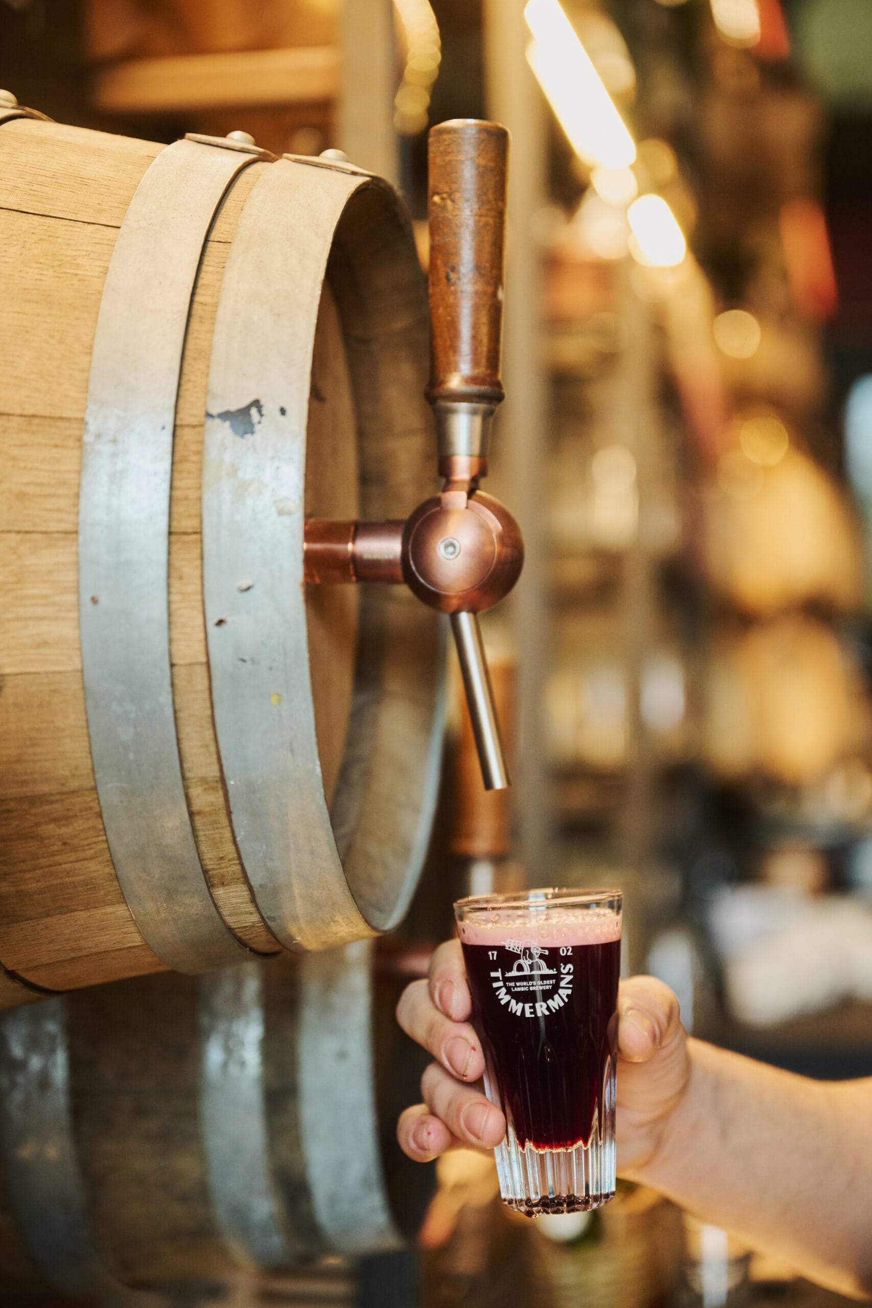 Een hand die een glas donker Timmermans bier vasthoudt onder een houten vat met een koperen tapkraan