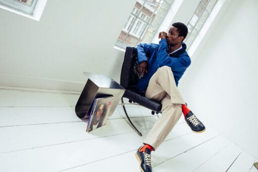 Zittende man in blauw jack, beige broek en donkere sneakers met oranje sokken in een lichte kamer