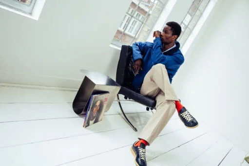 Zittende man in blauw jack, beige broek en donkere sneakers met oranje sokken in een lichte kamer