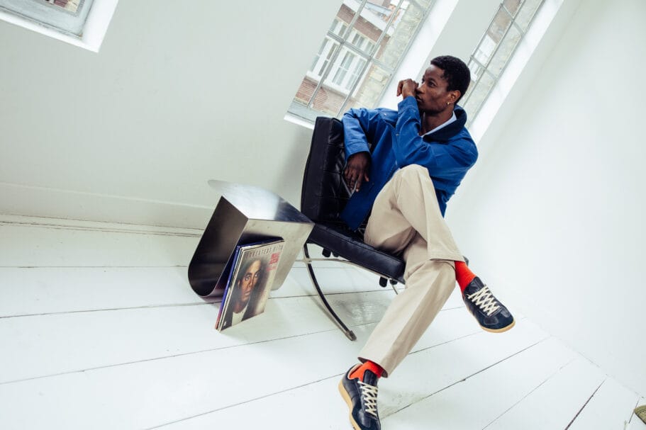Zittende man in blauw jack, beige broek en donkere sneakers met oranje sokken in een lichte kamer