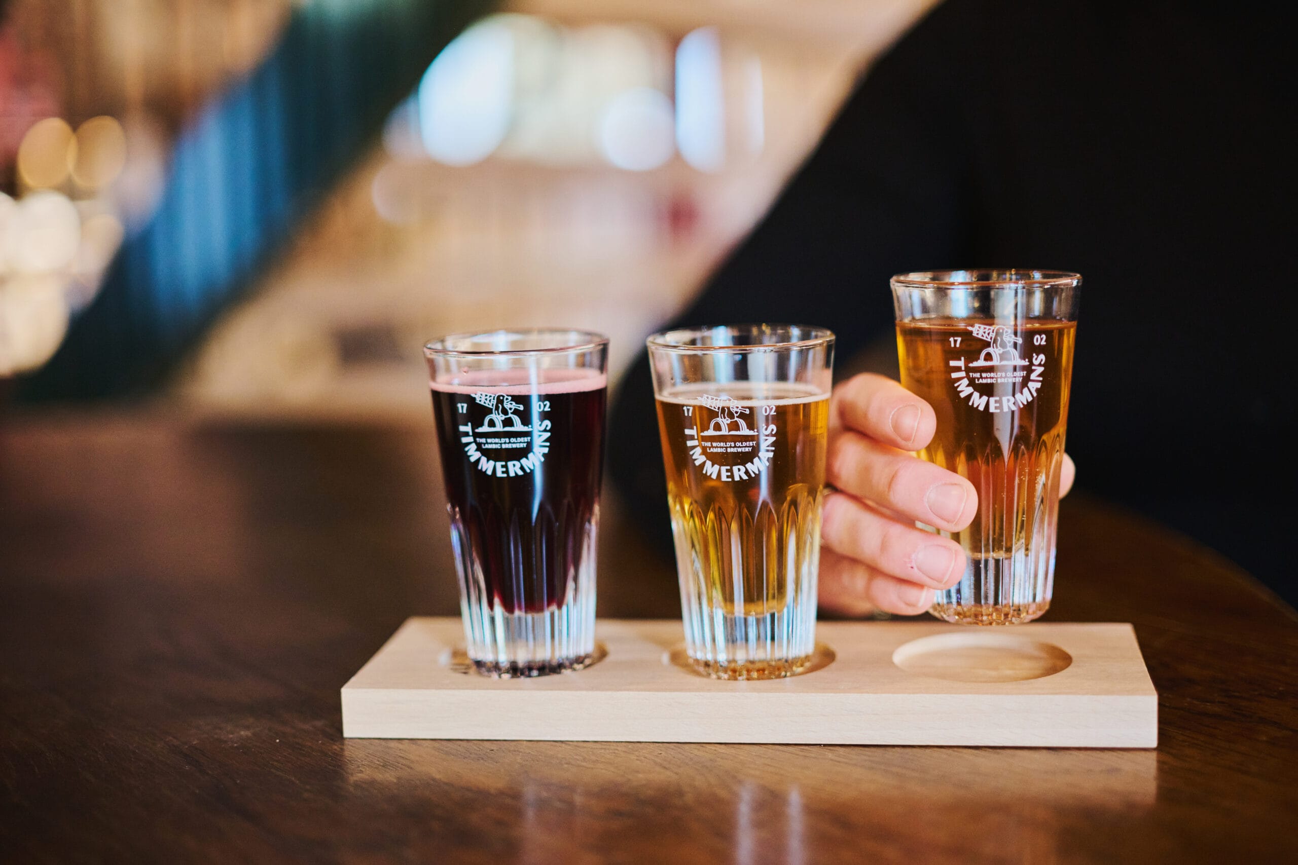 Drie proefglazen Timmermans lambiekbier in verschillende kleuren op een houten plankje