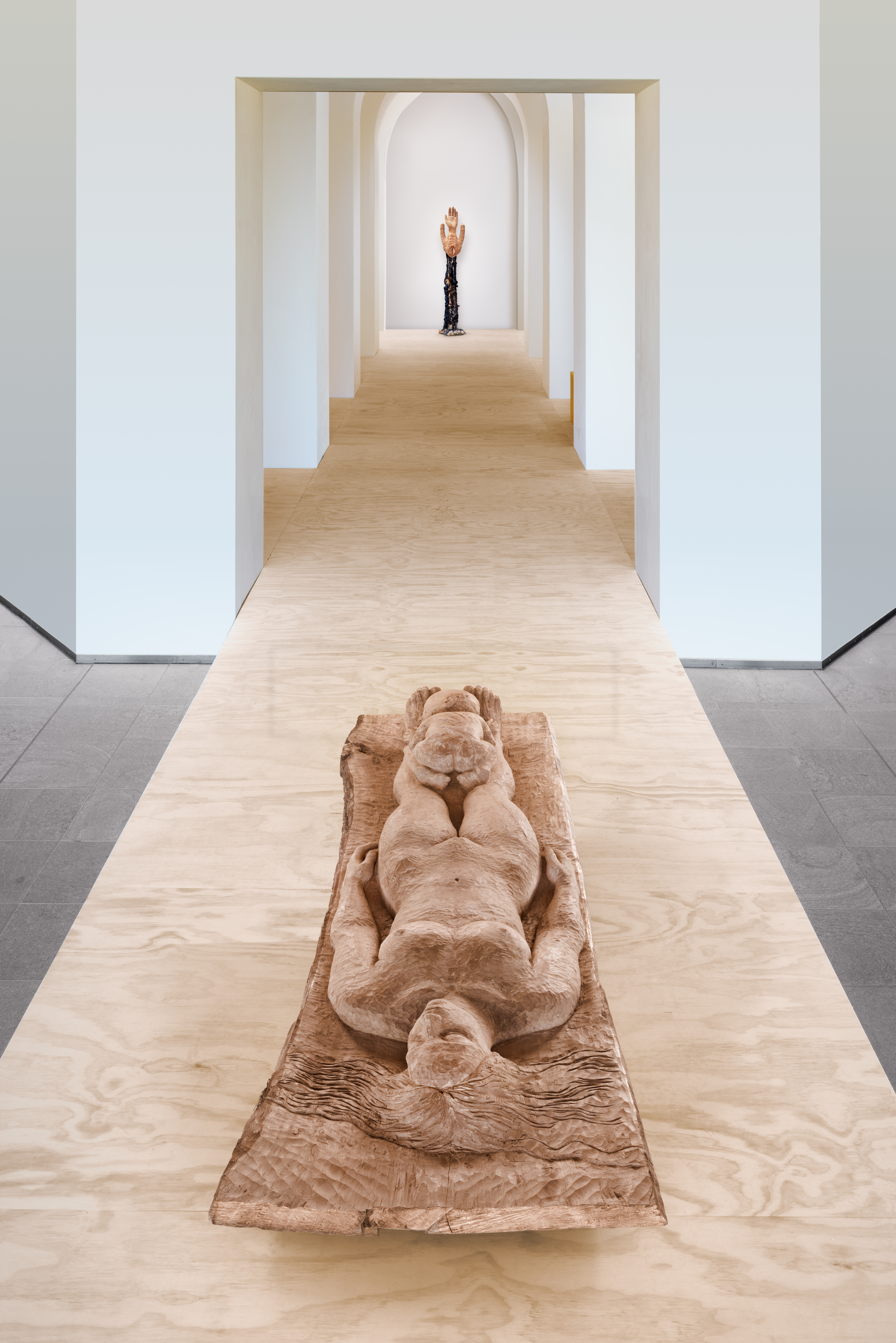 Houten sculptuur van een liggende vrouw op een verhoogd pad in een museumzaal, met in de verte een tweede kunstwerk, allemaal van de tentoonstelling Birthland van kunstenaar Femmy Otten in Museum Beelden aan Zee, Den Haag