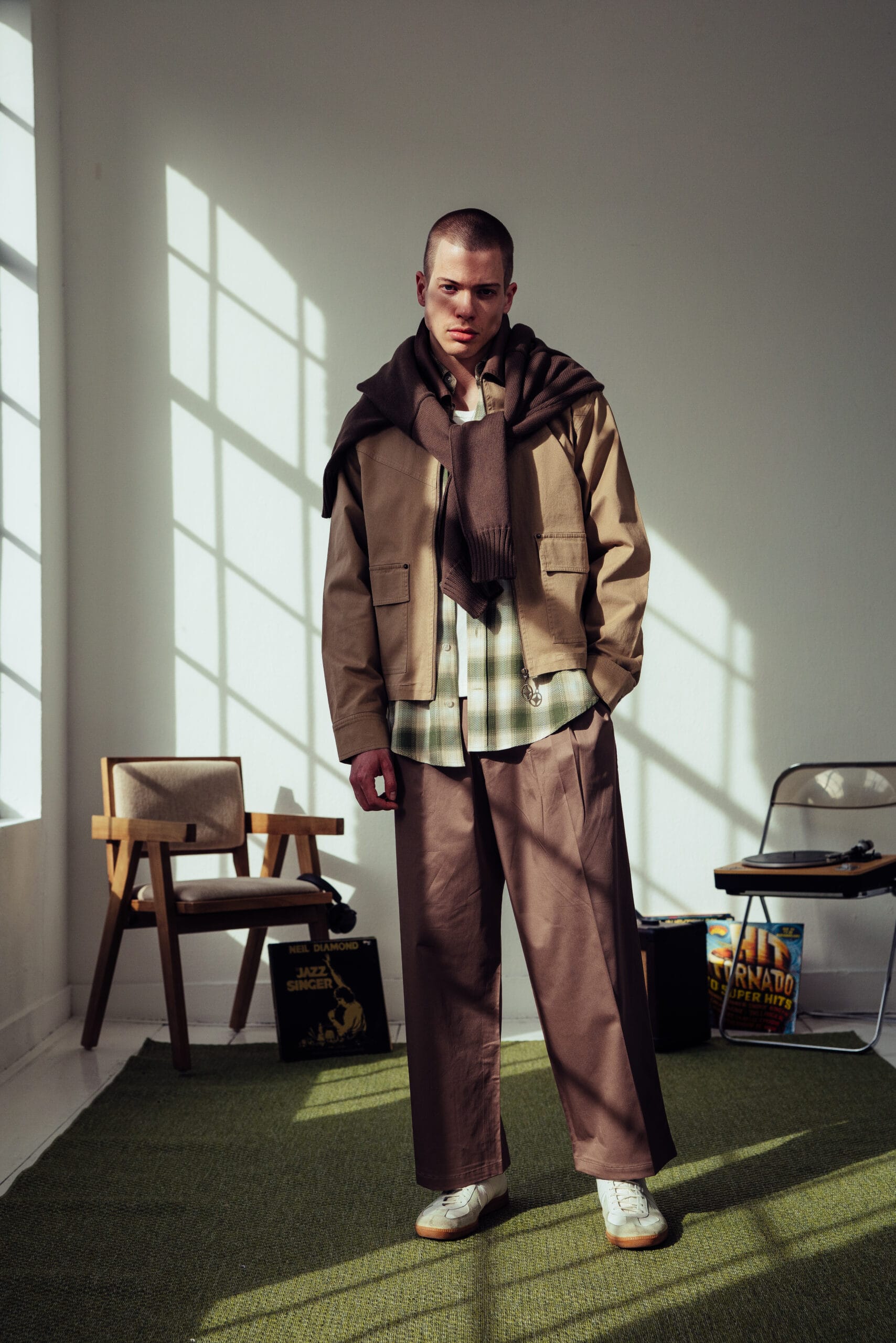 Man in een beige jasje, geruit overhemd en bruine pantalon met een trui over zijn schouders geknoopt