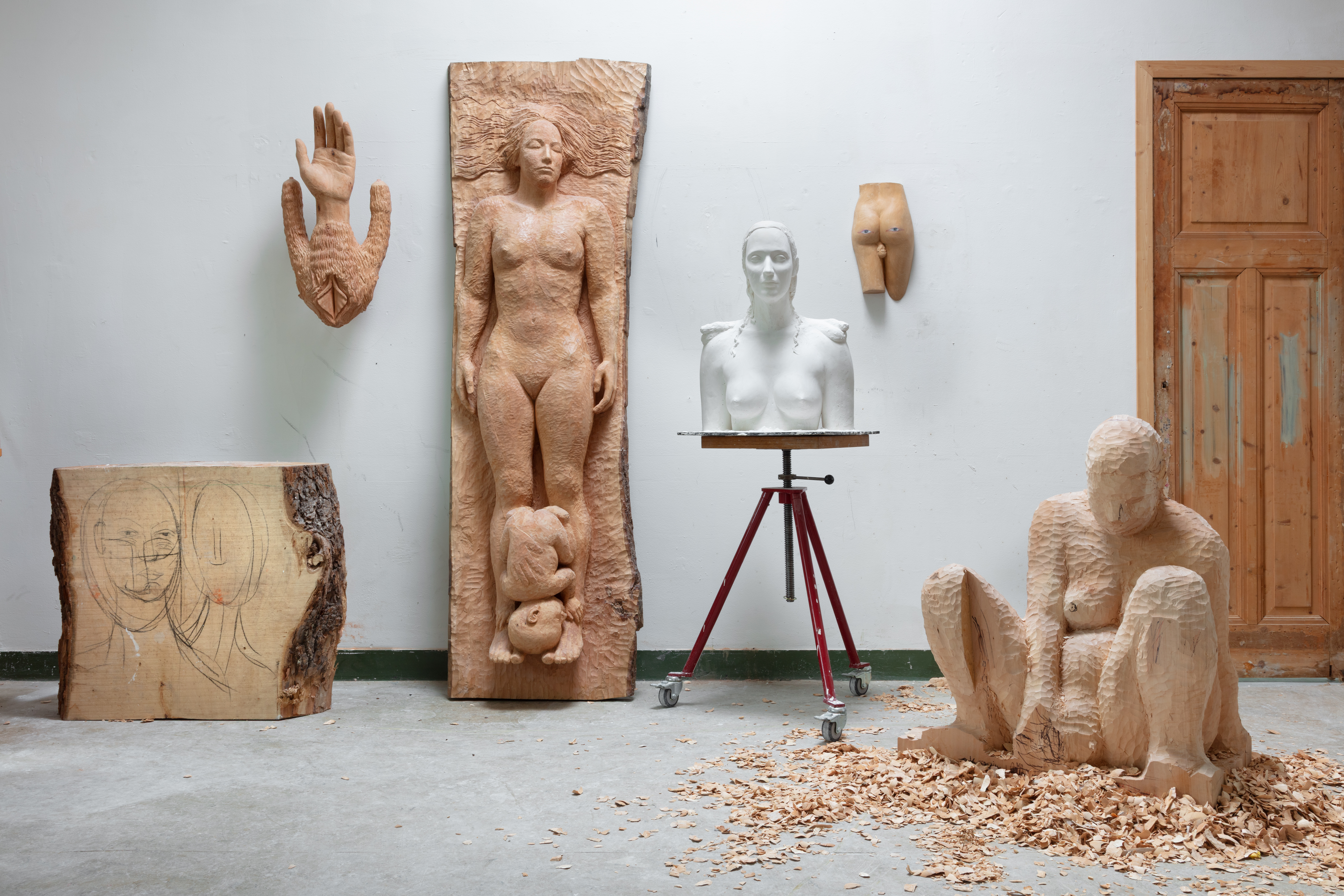 Verschillende houten en gipsen sculpturen van menselijke figuren in het atelier van kunstenaar Femmy Otten, met houtkrullen op de vloer