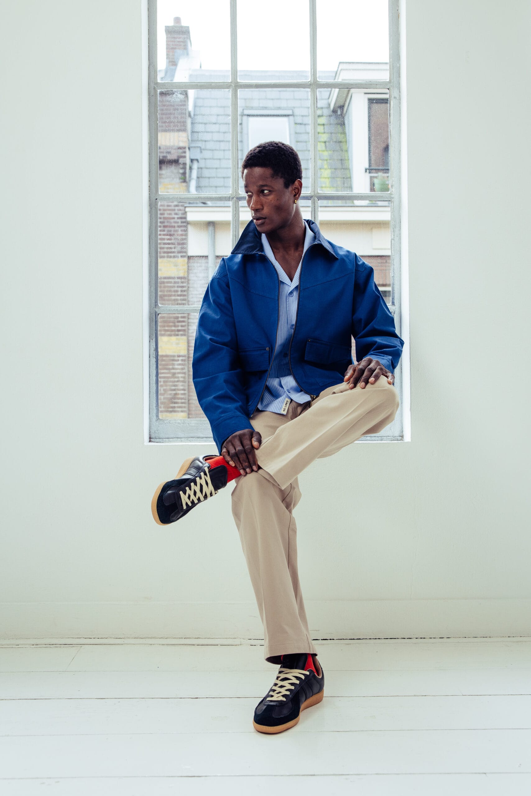 Een man zittend voor een raam in een blauw jack, gestreept overhemd, beige broek en donkere sneakers met rode accenten