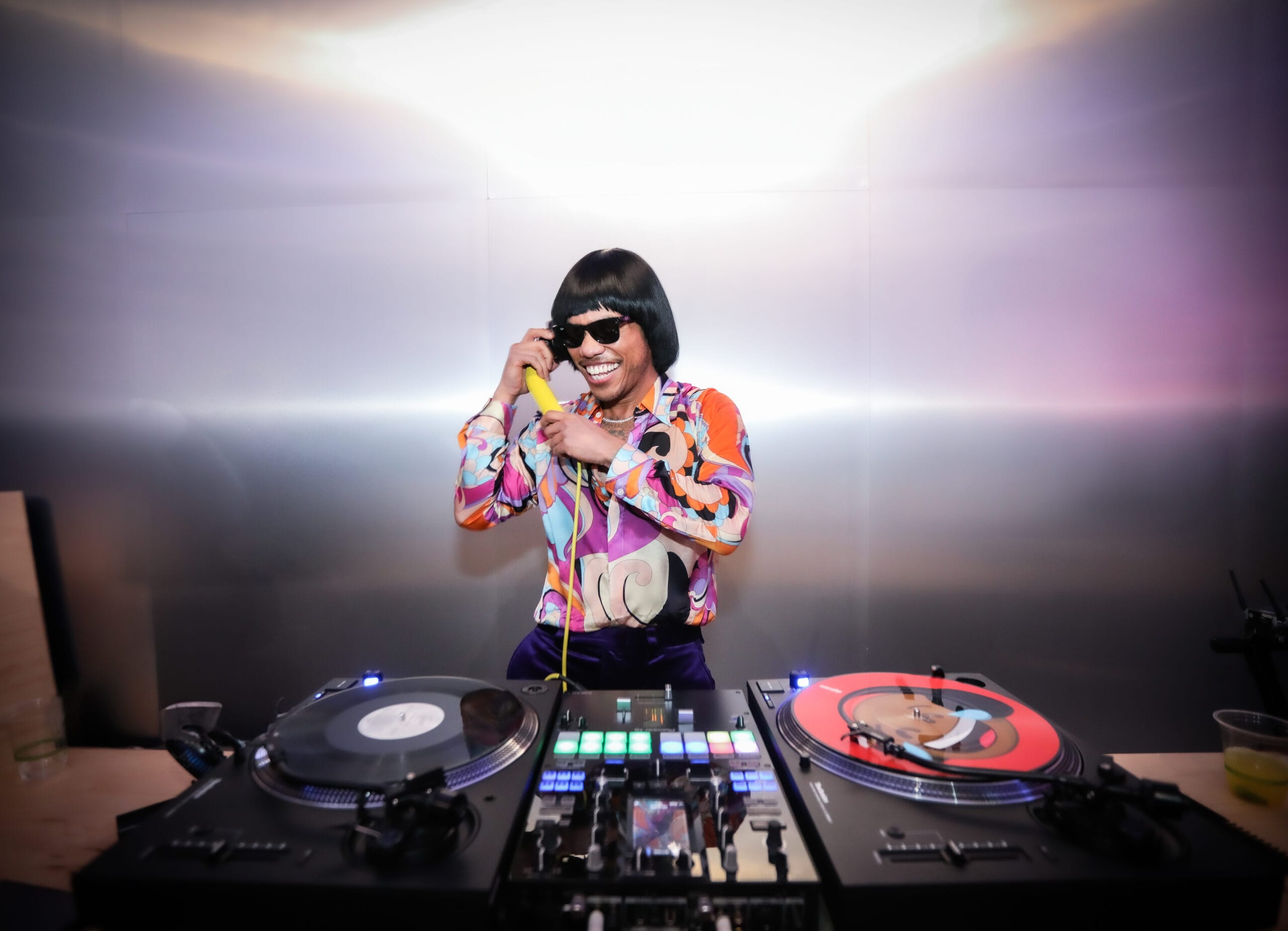 Anderson .Paak als DJ Pee .Wee met een pruik en zonnebril, lachend achter een mengpaneel en draaitafels