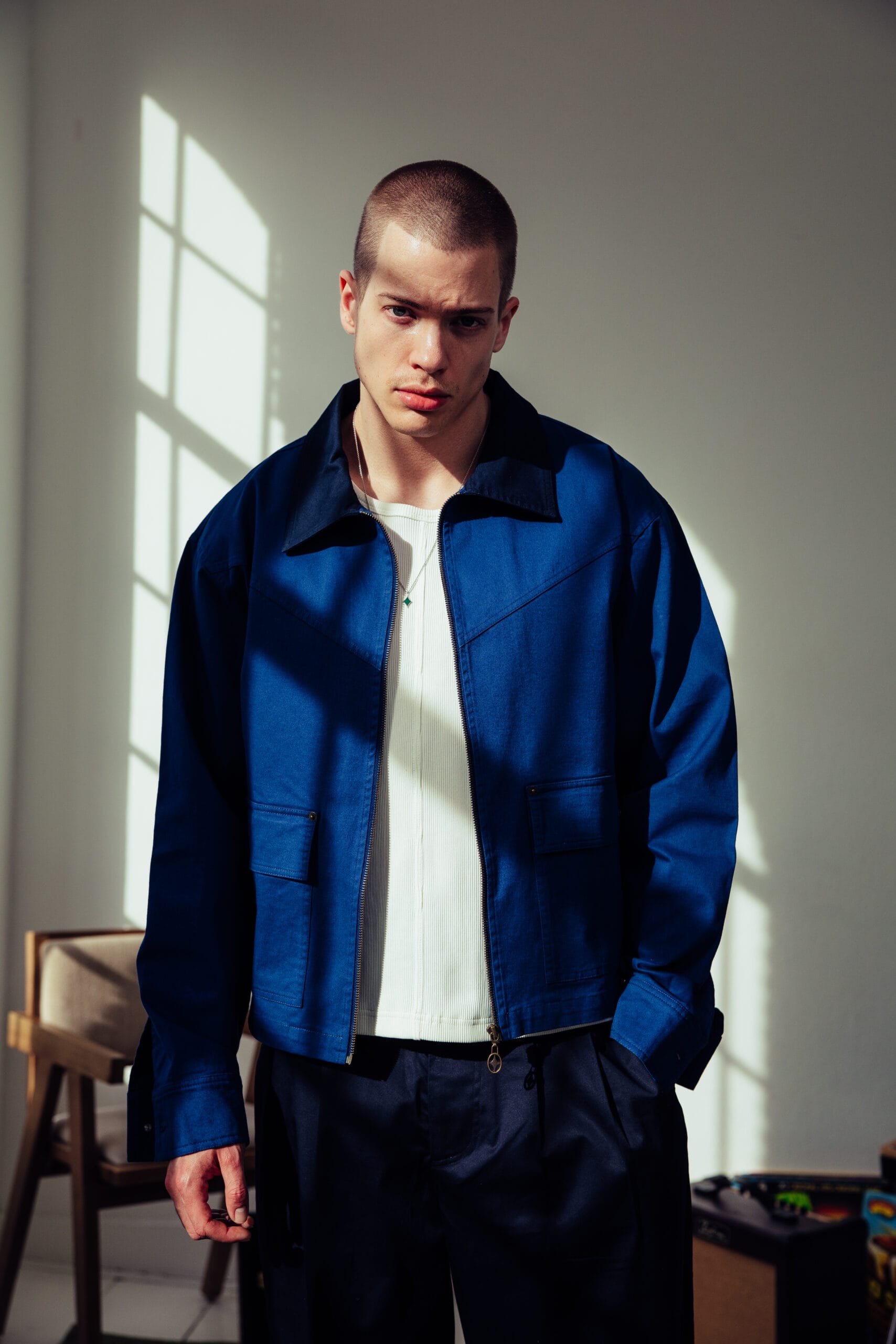 Een man met kortgeschoren haar draagt een blauw jack over een wit shirt en een donkere broek in een zonverlichte kamer