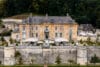 Vooraanzicht van het historisch landhuis Chateau Neercanne van gele mergelsteen met een terras en witte parasols tegen een beboste heuvel