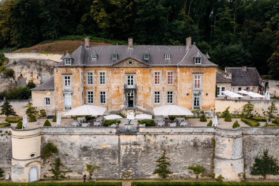 Vooraanzicht van het historisch landhuis Chateau Neercanne van gele mergelsteen met een terras en witte parasols tegen een beboste heuvel
