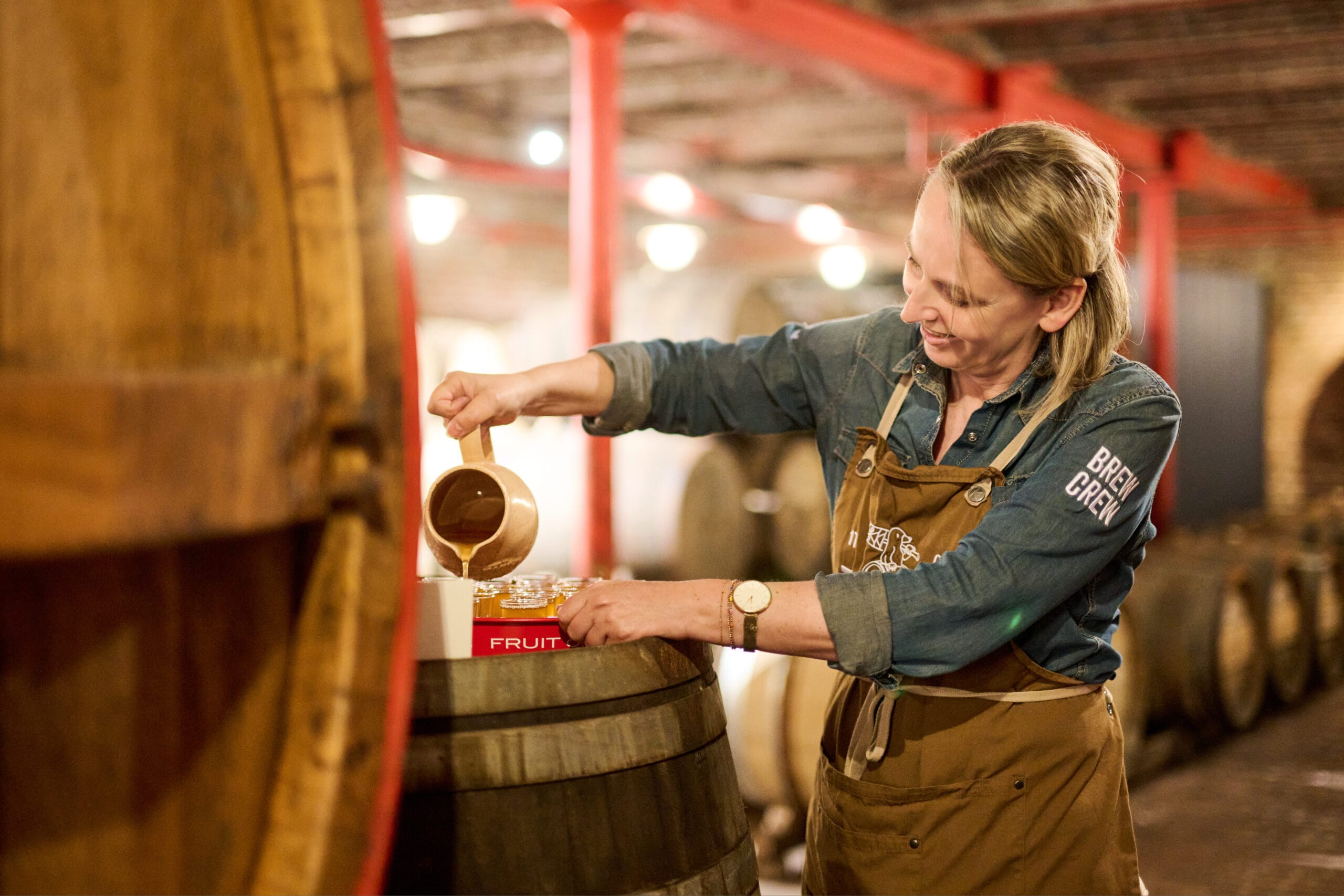 Een glimlachende vrouw van de Brew Crew schenkt vloeistof uit een kan in glazen op een houten vat in een brouwerij