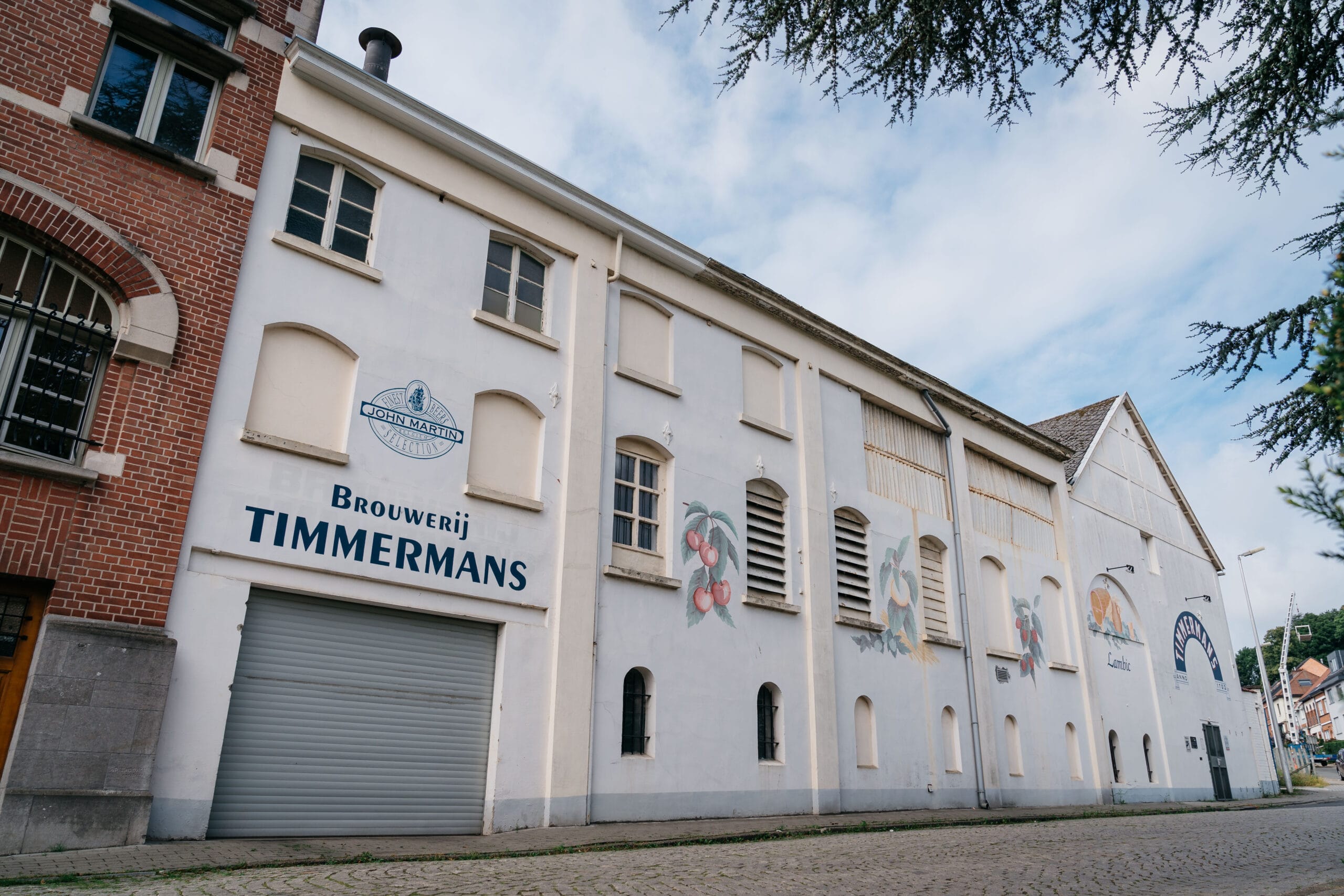 De witte gevel van Brouwerij Timmermans met muurschilderingen van fruit en het logo van John Martin