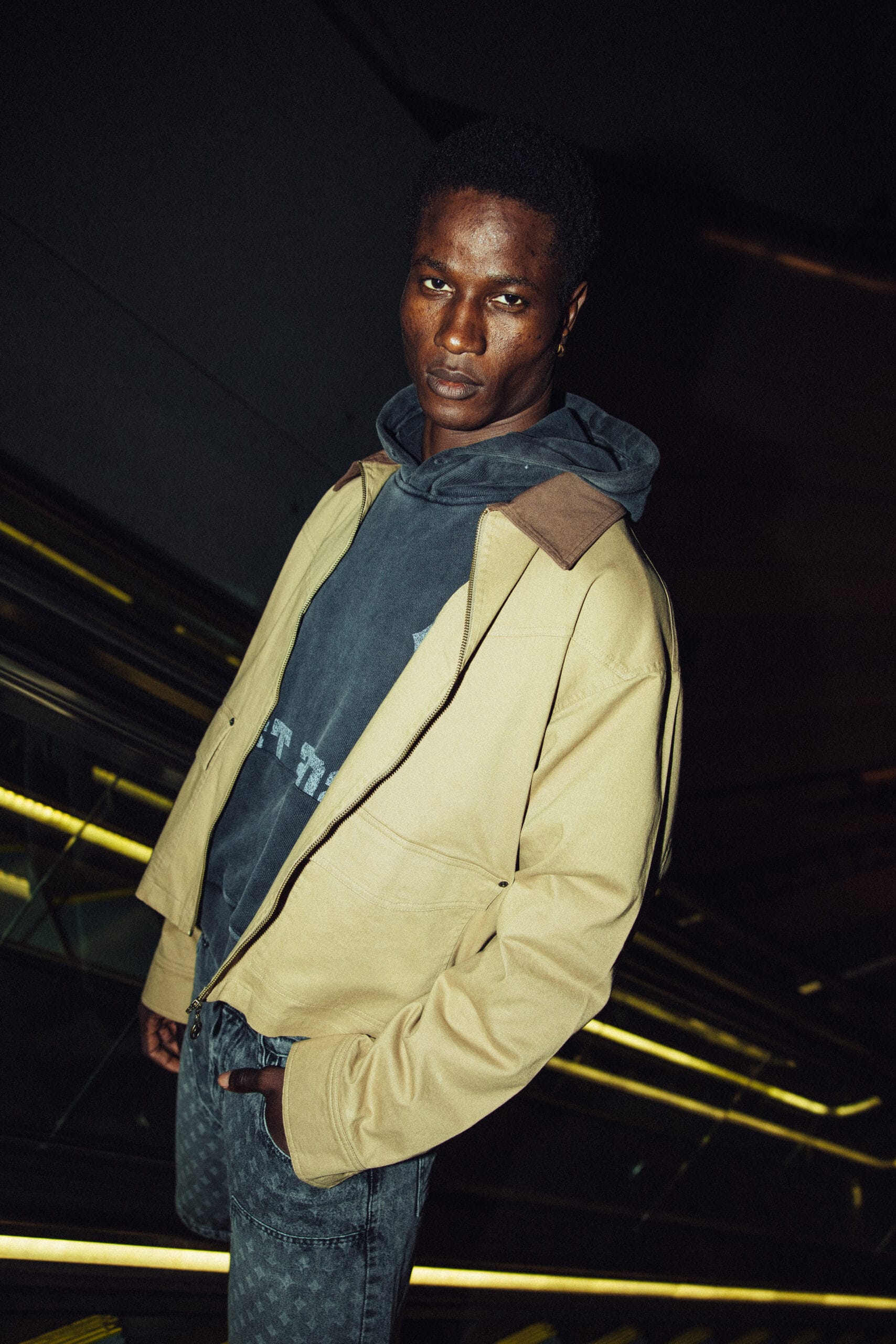 Man met een donkere huidskleur draagt een beige jack over een grijze hoodie en een donkere jeans
