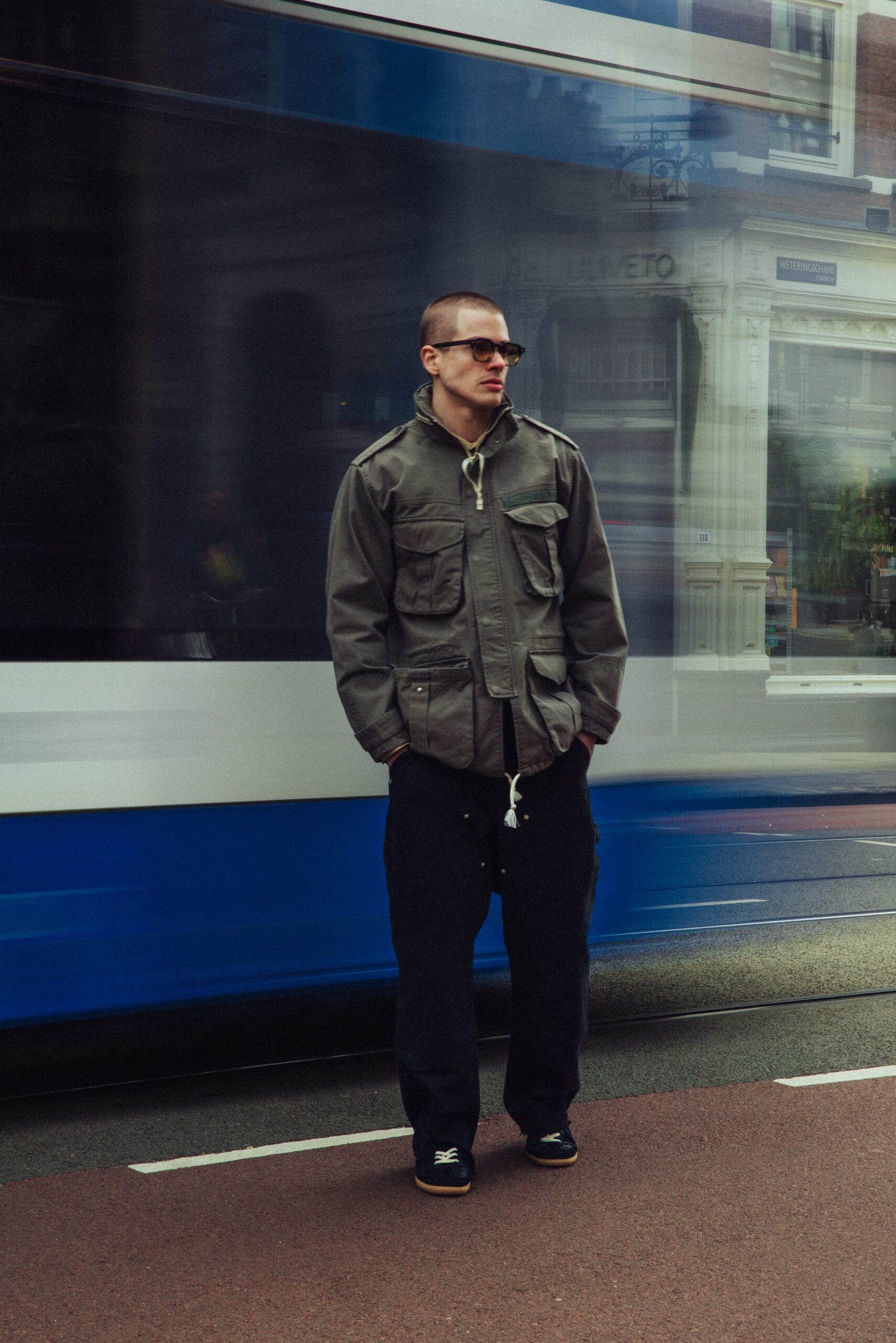Een man in een olijfgroen veldjack en donkere broek staat op een tramhalte terwijl een blauw-witte tram voorbijrijdt