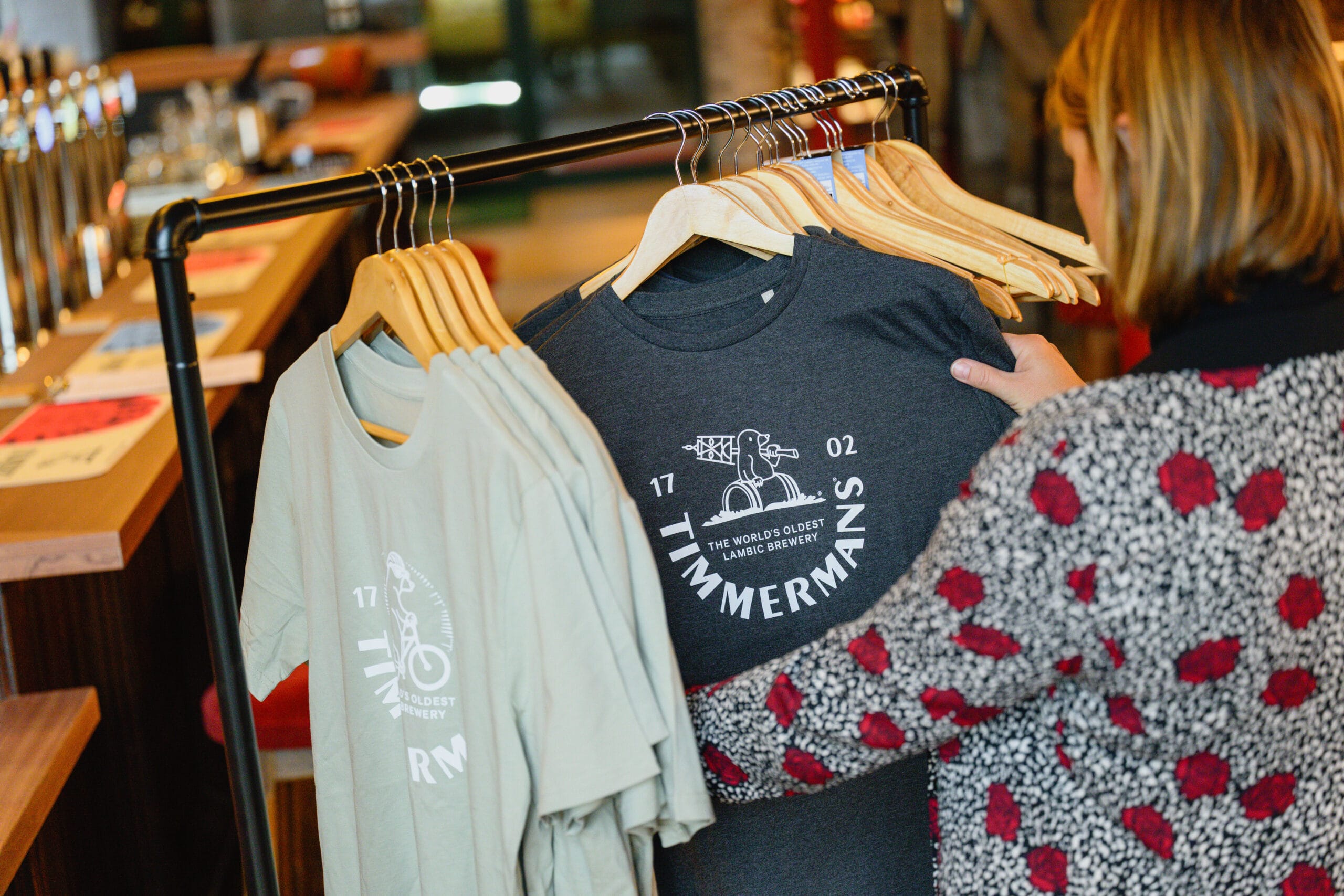 Een vrouw bekijkt T-shirts van brouwerij Timmermans aan een kledingrek in een café