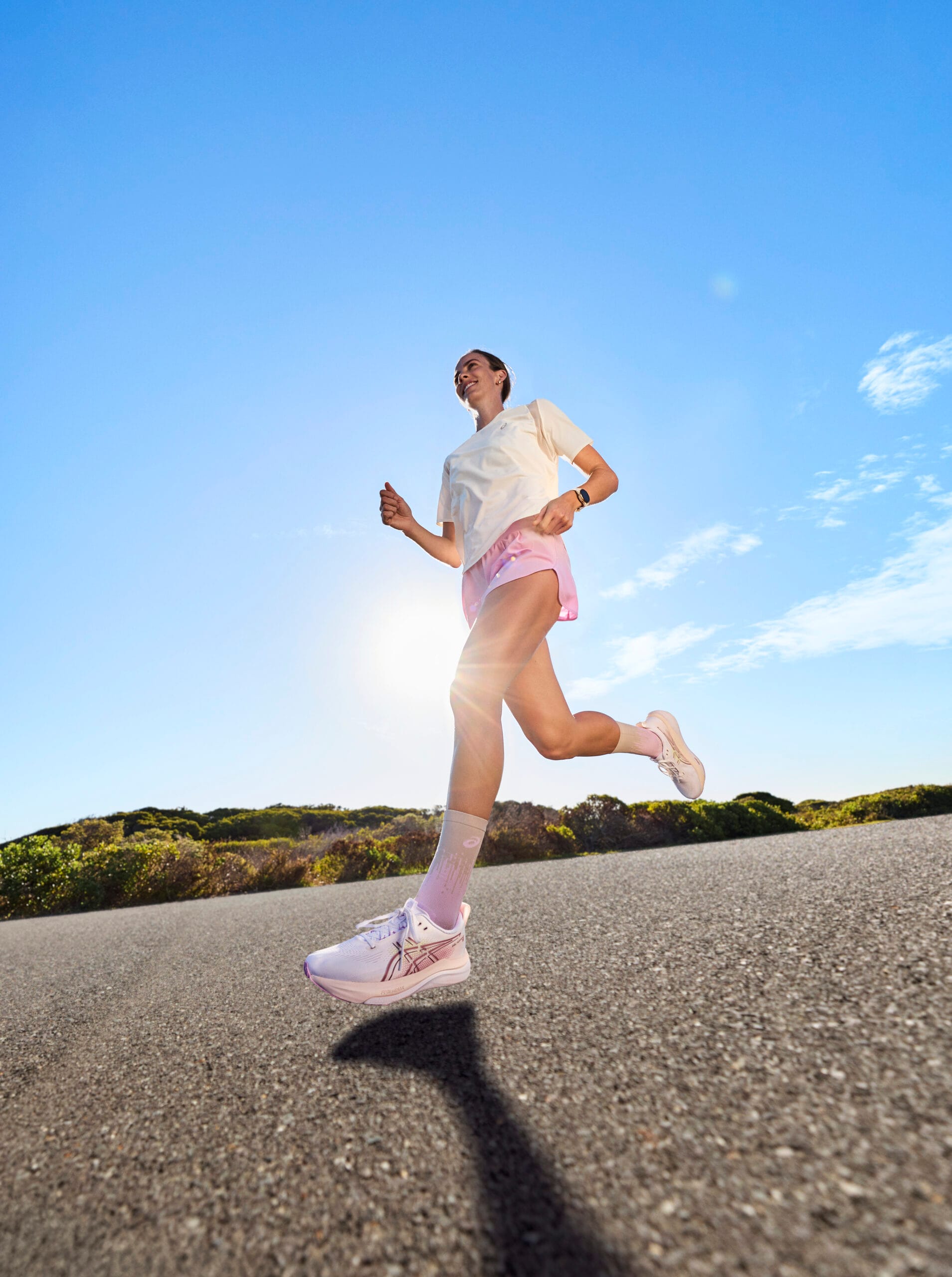 Een lachende vrouw hardlopend op de ASICS GEL-KAYANO 33 op een asfaltweg tegen een helderblauwe lucht bij zonsopgang
