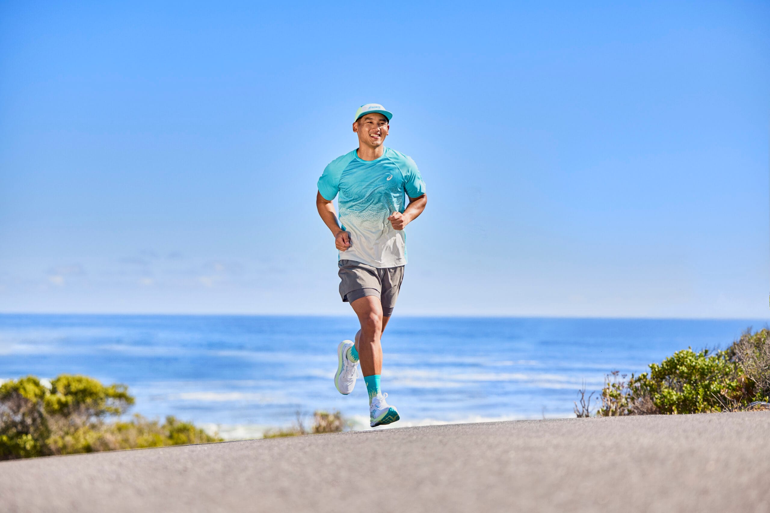 Glimlachende man in sportkleding hardlopend op de ASICS GEL-KAYANO 33 op een weg langs de kust met de oceaan op de achtergrond