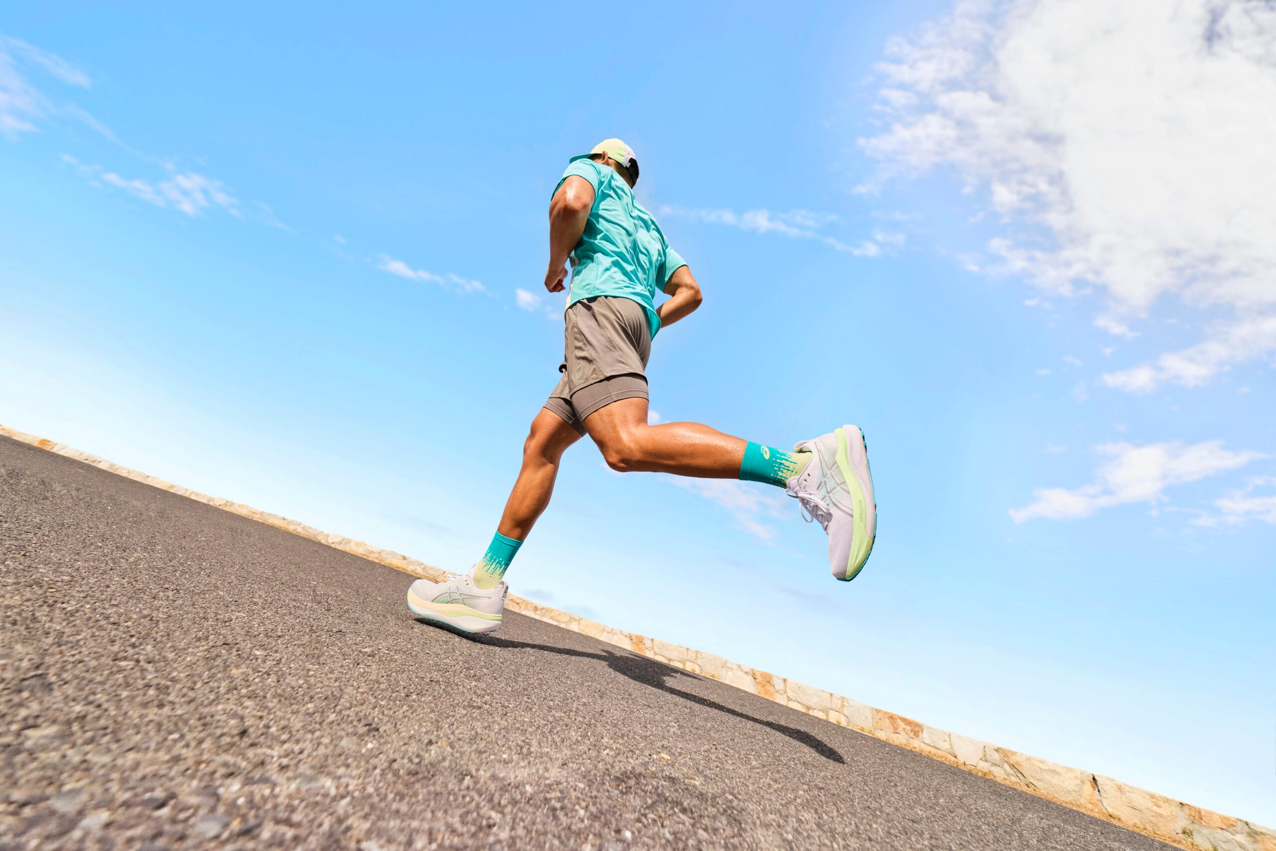 Een hardloper in sportkleding van ASICS rent op de GEL-KAYANO 33 over een geasfalteerde weg tegen een helderblauwe lucht
