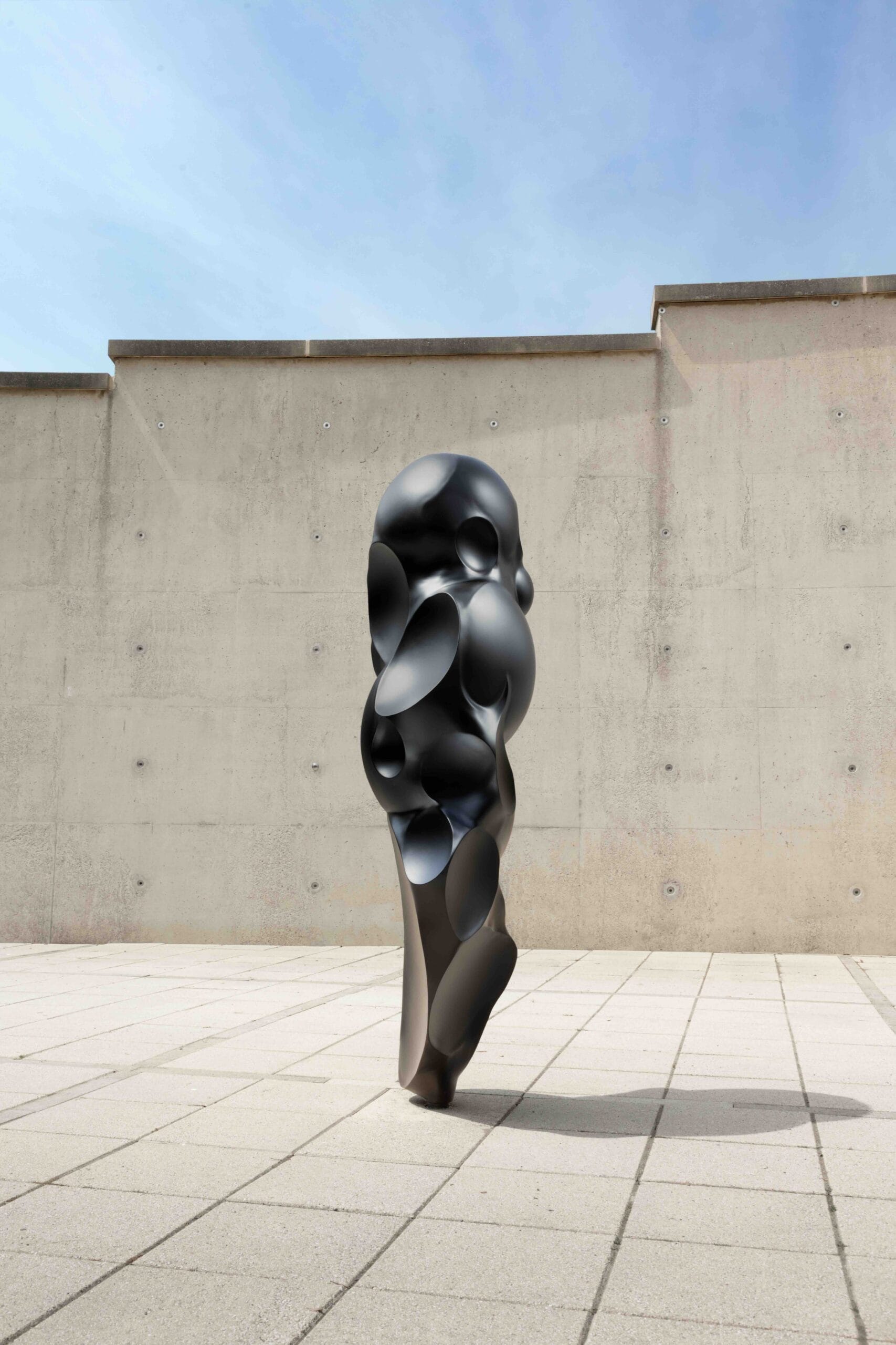 Een abstract, verticaal zwart beeldhouwwerk met organische vormen en ronde inkepingen van de tentoonstelling Rui Chafes X Alberto Giacometti. Gris Vide Cris III op een terras van Museum Beelden aan Zee