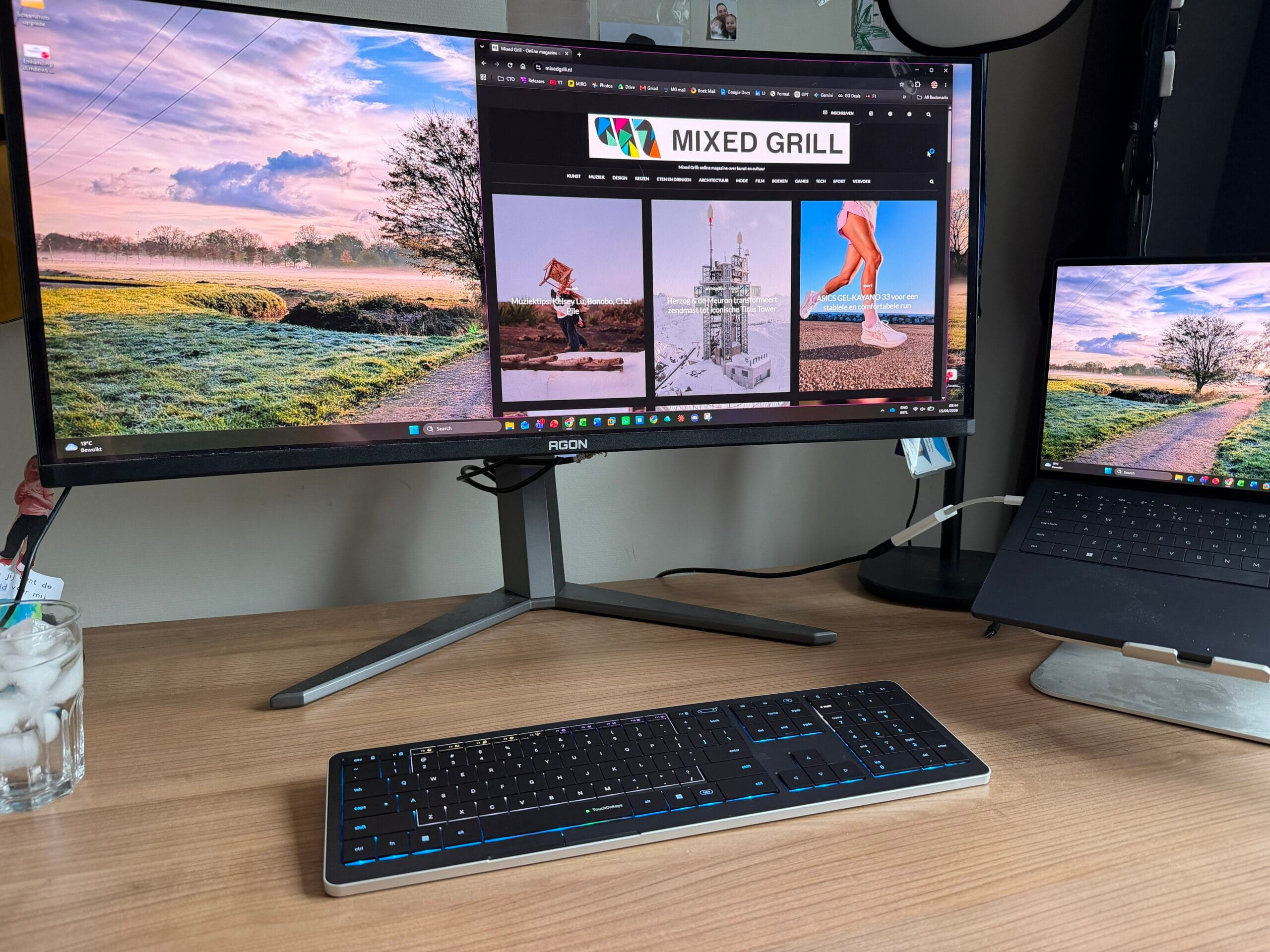 Een bureau met een breed computerscherm waarop de website van Mixed Grill openstaat, een verlicht toetsenbord en een laptop