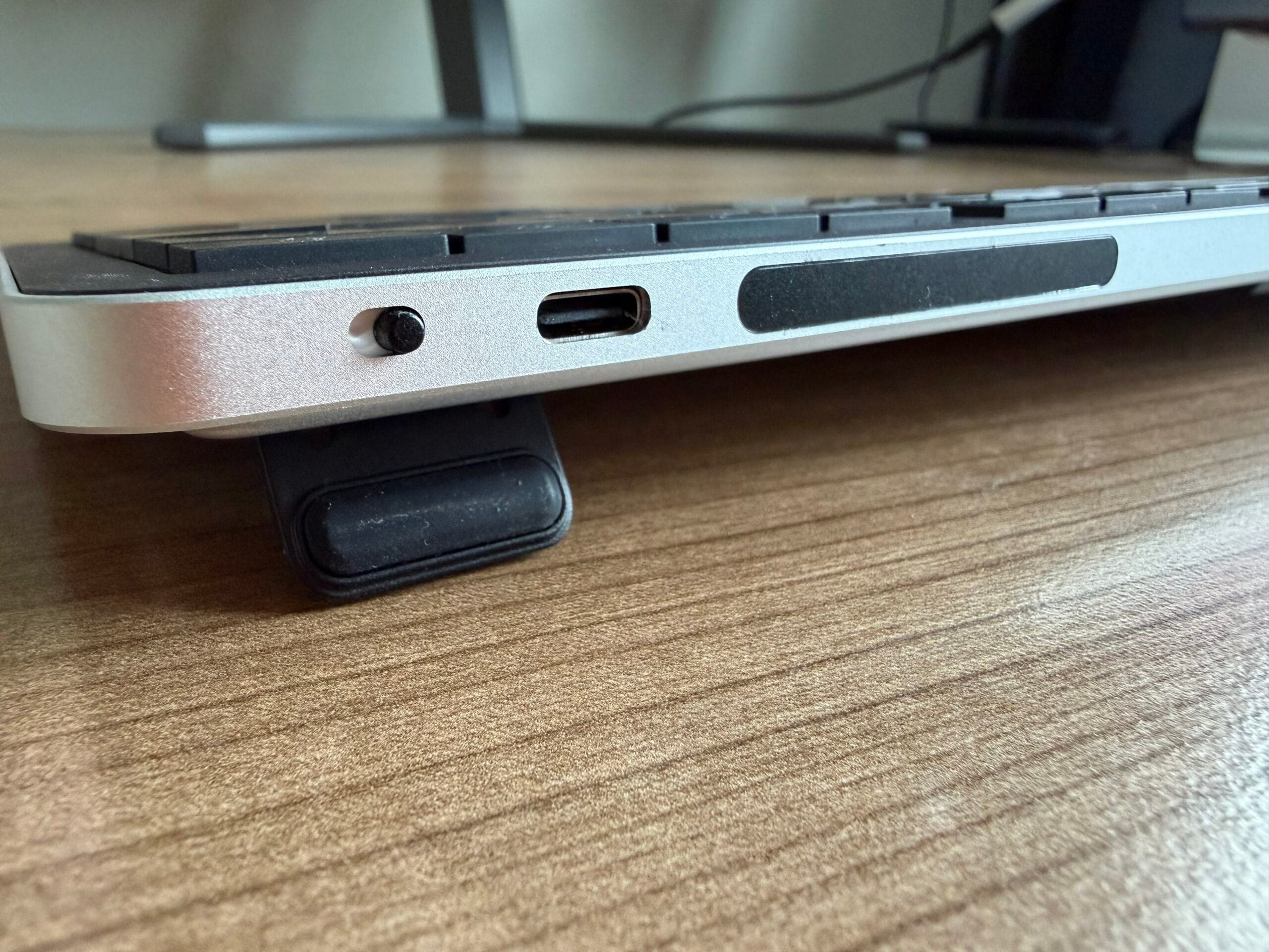 Zijaanzicht van een toetsenbord met een aan-uitknop, USB-C-poort en een uitgeklapt pootje op een houten bureau