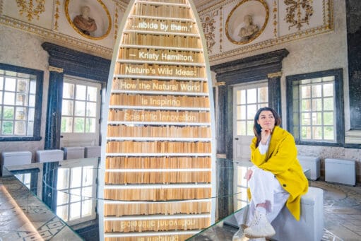 Een vrouw in een gele jas zit naast een kegelvormige boekenkast met een citaat van Kristy Hamilton over natuur en innovatie