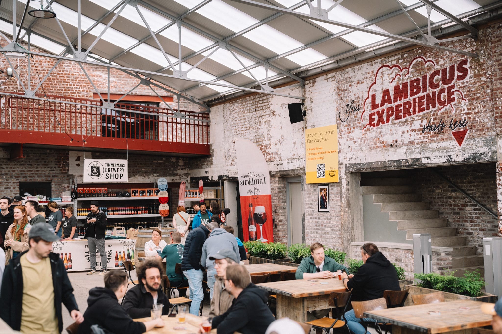 Mensen zitten aan houten tafels in de Lambicus Experience van Brouwerij Timmermans, met een winkel op de achtergrond