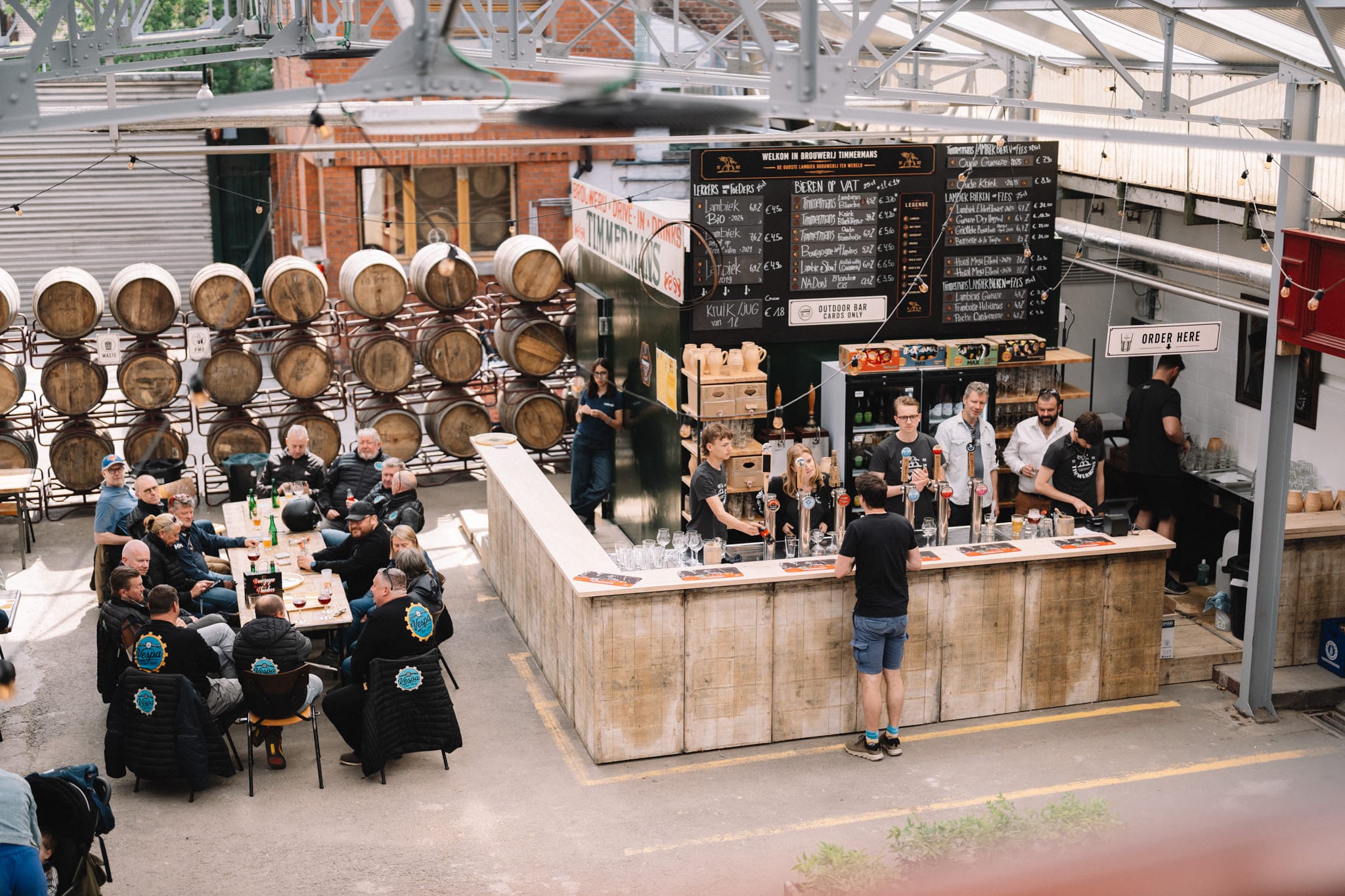 Gasten aan houten tafels en personeel achter een bar in de openluchtbrouwerij van Timmermans, met bieraten op de achtergrond