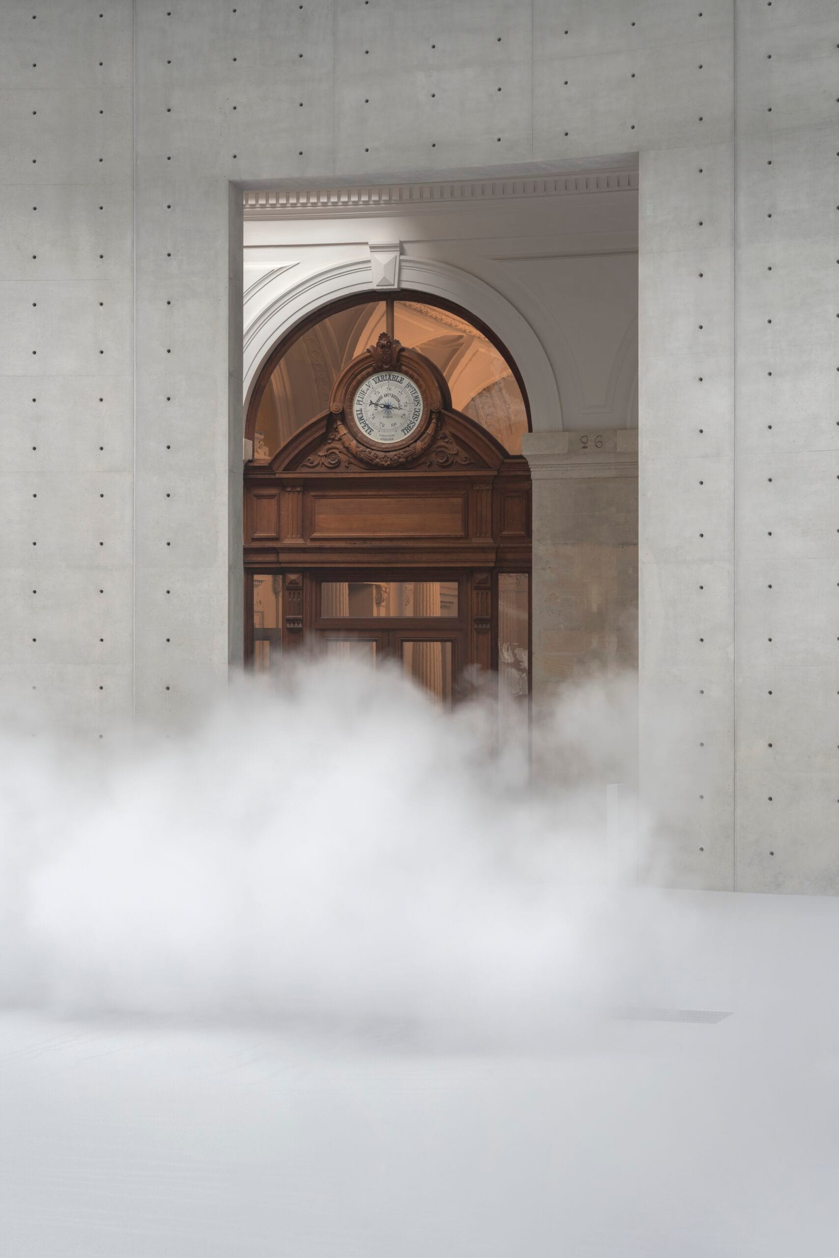 Een dichte mistwolk op een binnenplaats voor een klassieke houten deuropening met een barometer in een modern betonnen kader