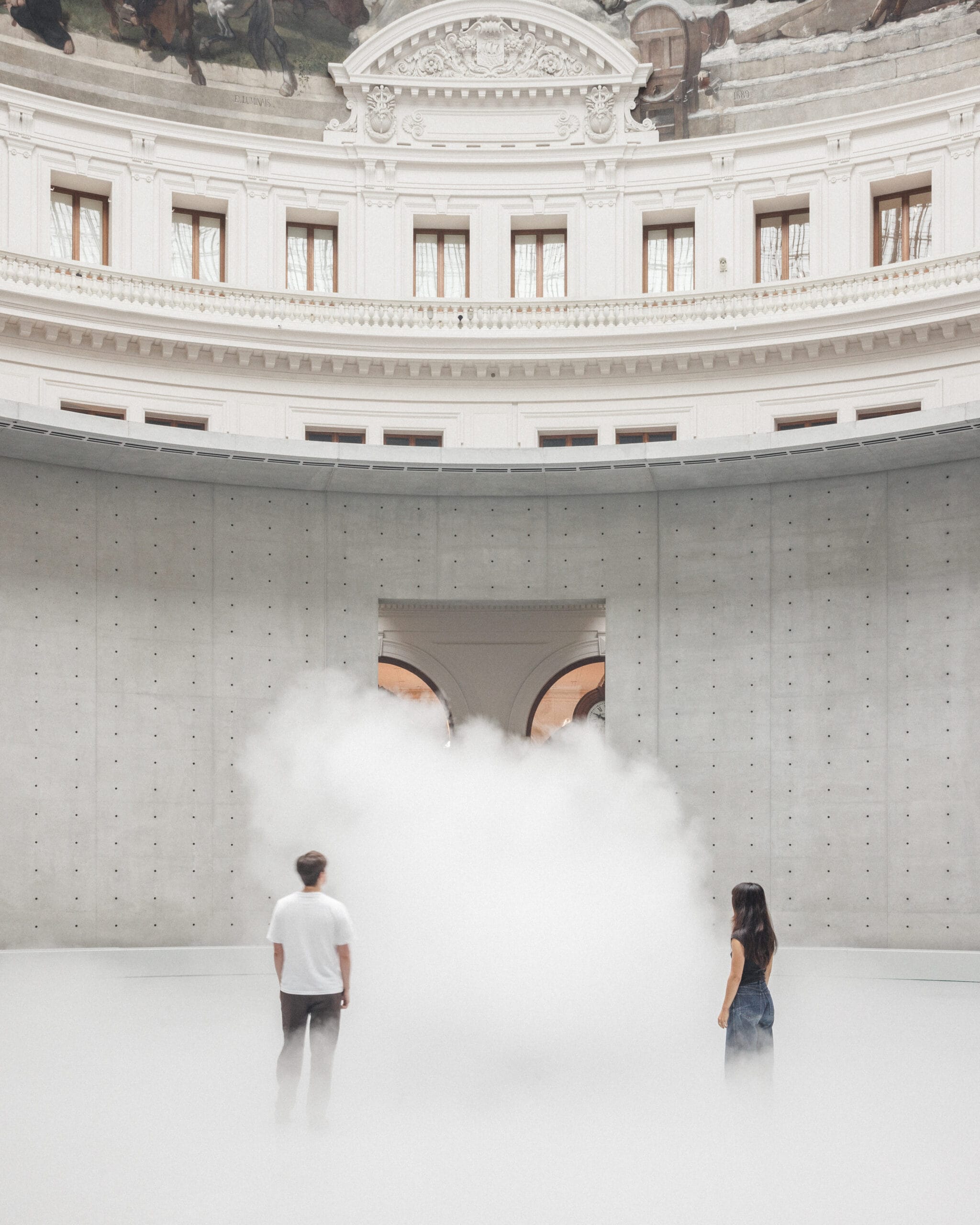 Twee mensen staan in een grote, ronde zaal met een betonnen wand en een witte mistwolk in het midden
