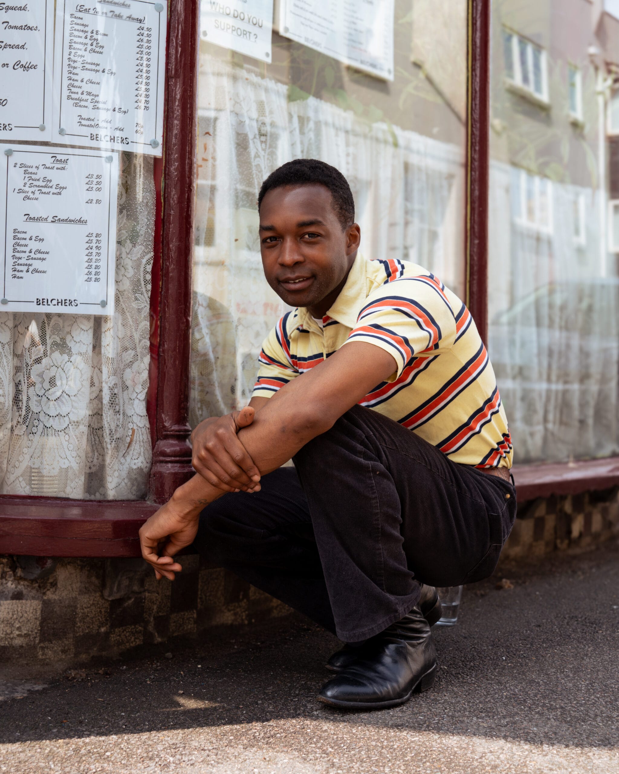 zanger Jalen Ngonda in een gestreept poloshirt en corduroy broek hurkt voor de etalage van een café met menukaarten