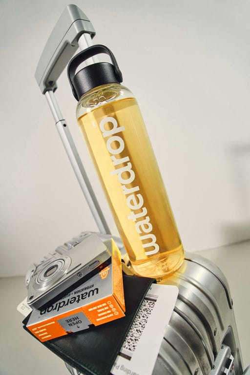 Een Waterdrop-fles met geel drankje op een koffer naast een camera, paspoort en een doosje Waterdrop-hydratatieblokjes