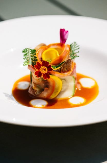Een verfijnd gerecht van opgerolde groenten geserveerd in een oranje saus met witte druppels, gegarneerd met eetbare bloemen, bij Les Salons op Château St. Gerlach 