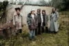 Vijf mannen van de band Swim deep in winterjassen en mutsen poseren voor een golfplaten wand in een landelijke omgeving