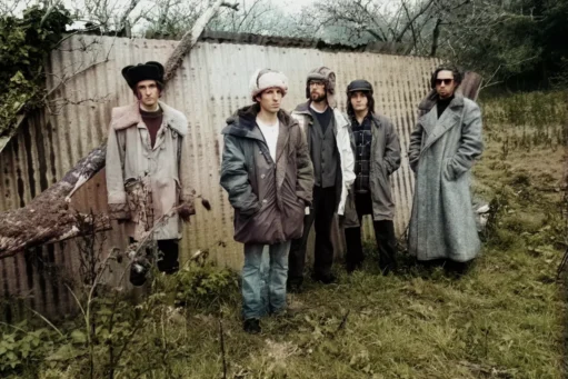 Vijf mannen van de band Swim deep in winterjassen en mutsen poseren voor een golfplaten wand in een landelijke omgeving