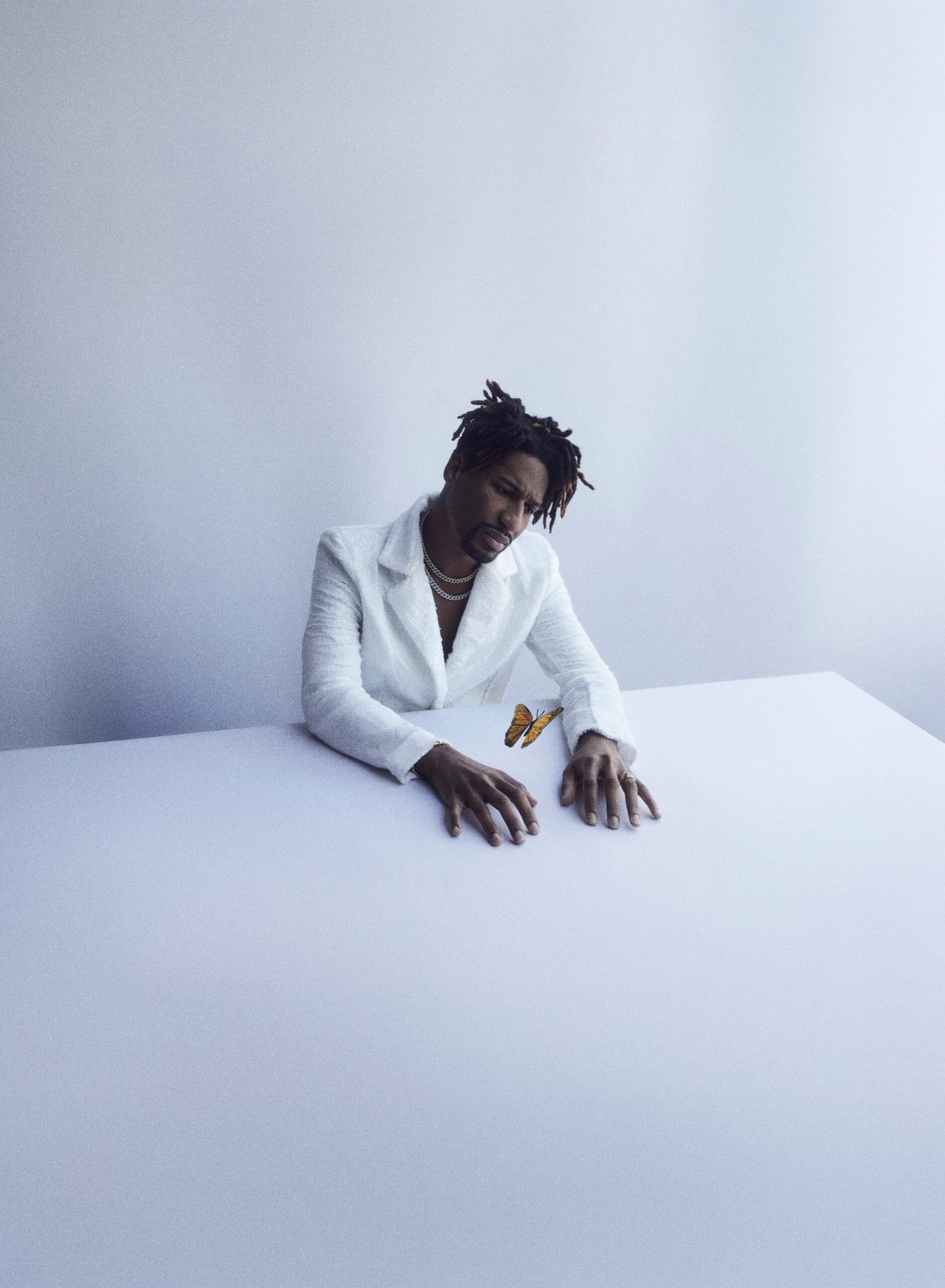 Jon Batiste in een wit pak aan een witte tafel, kijkend naar een monarchvlinder die tussen zijn handen vliegt