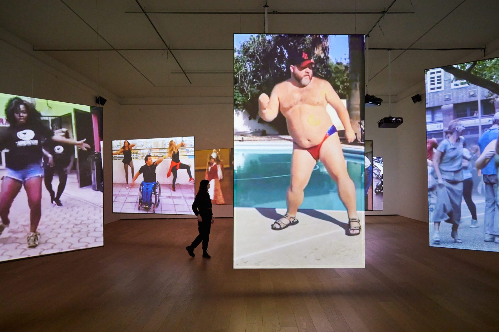 Een persoon loopt door een museumzaal met grote hangende schermen waarop diverse dansende mensen te zien zijn