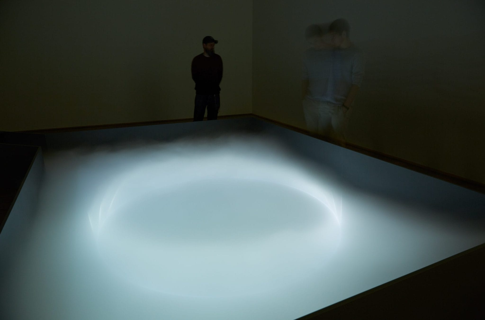 Een verlichte cirkel van mist in een donkere expositieruimte met toeschouwers op de achtergrond