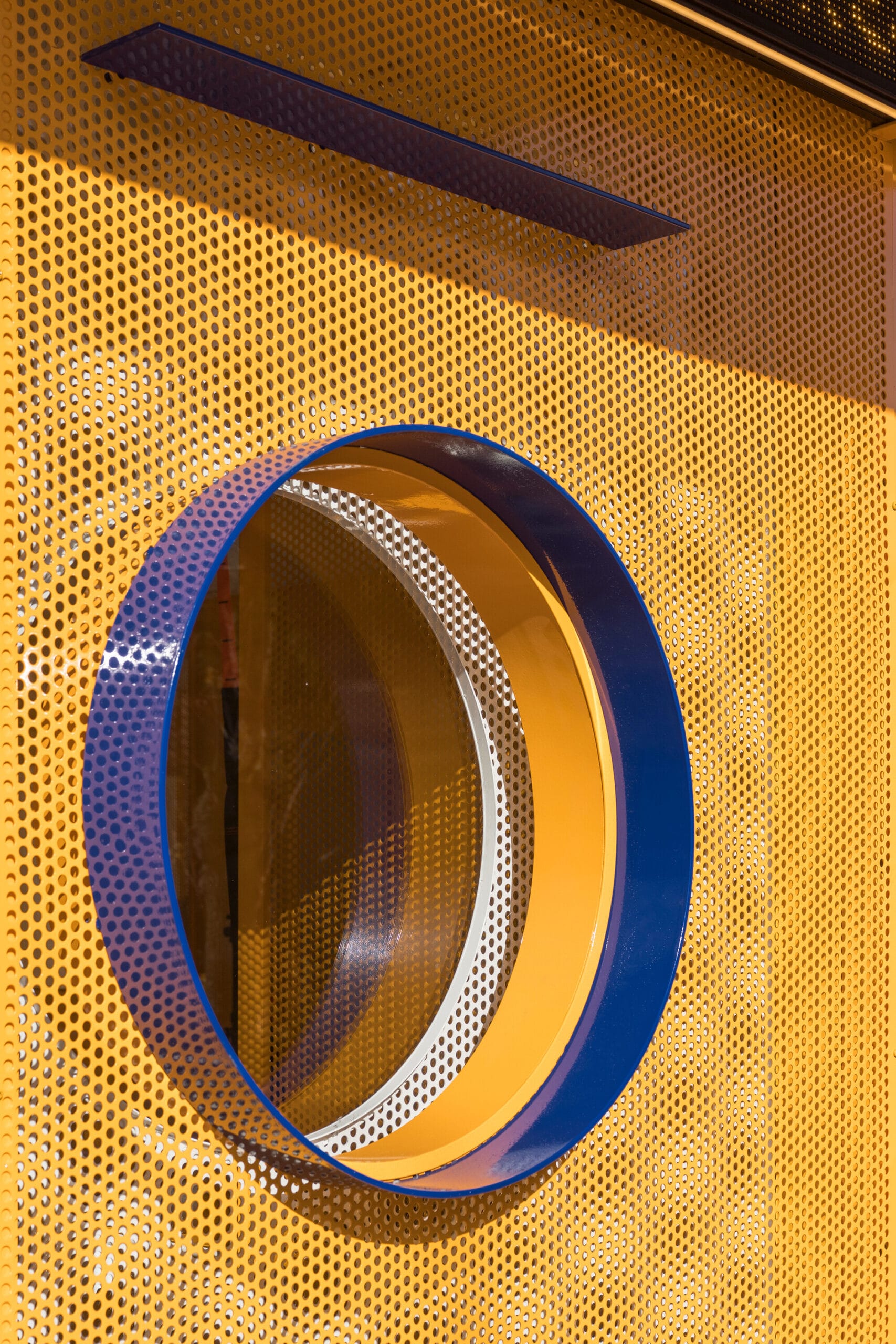 Een rond raam met een blauwe en gele omlijsting in een gele geperforeerde metalen wand