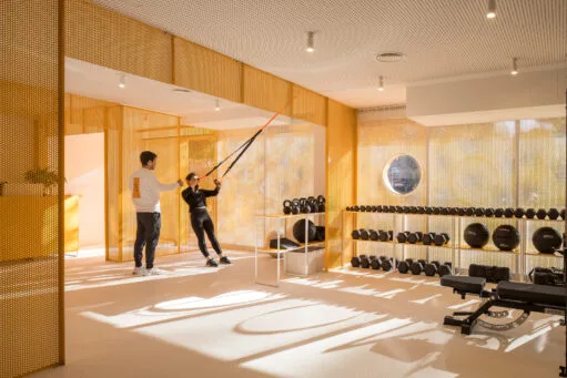 Een vrouw traint met TRX-banden onder begeleiding van een coach in een moderne sportschool met gele geperforeerde wanden