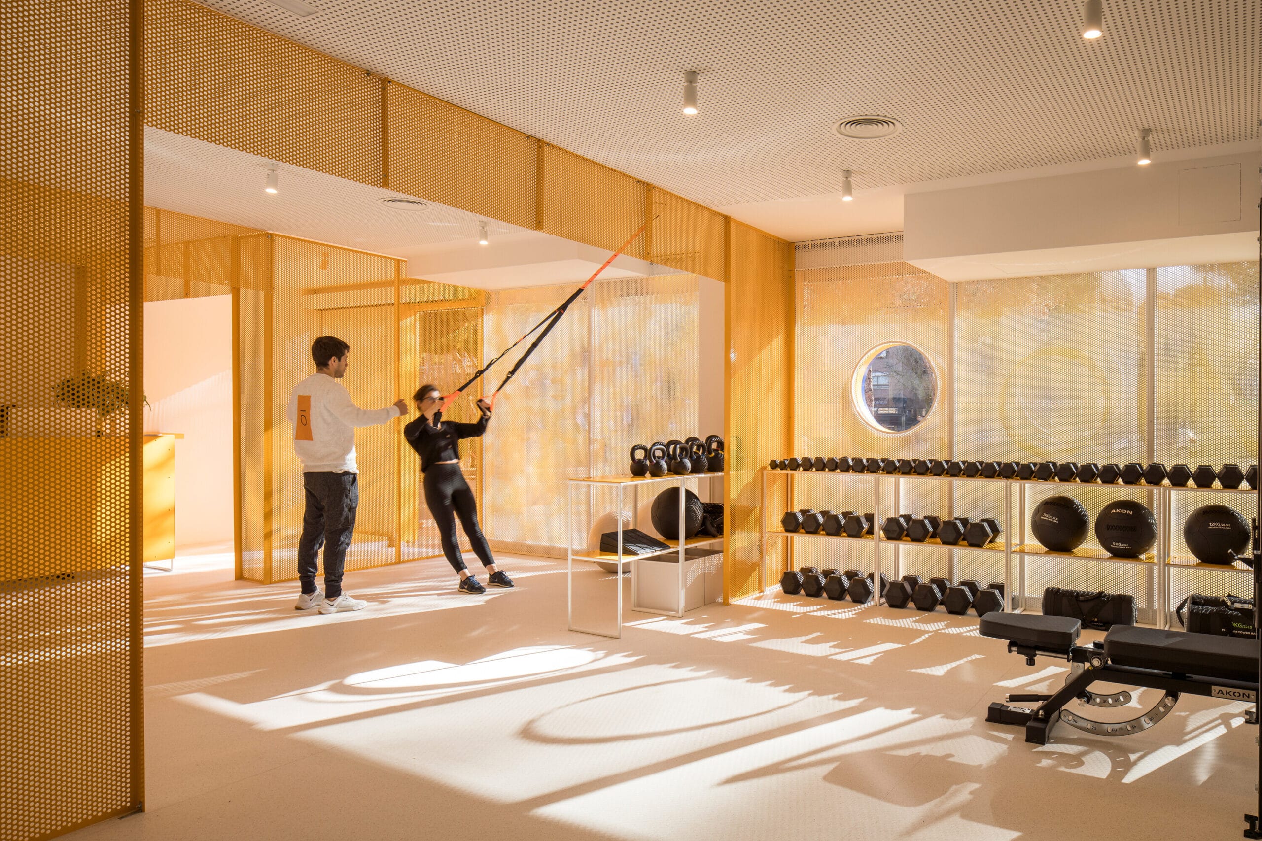 Een vrouw traint met TRX-banden onder begeleiding van een coach in een moderne sportschool met gele geperforeerde wanden