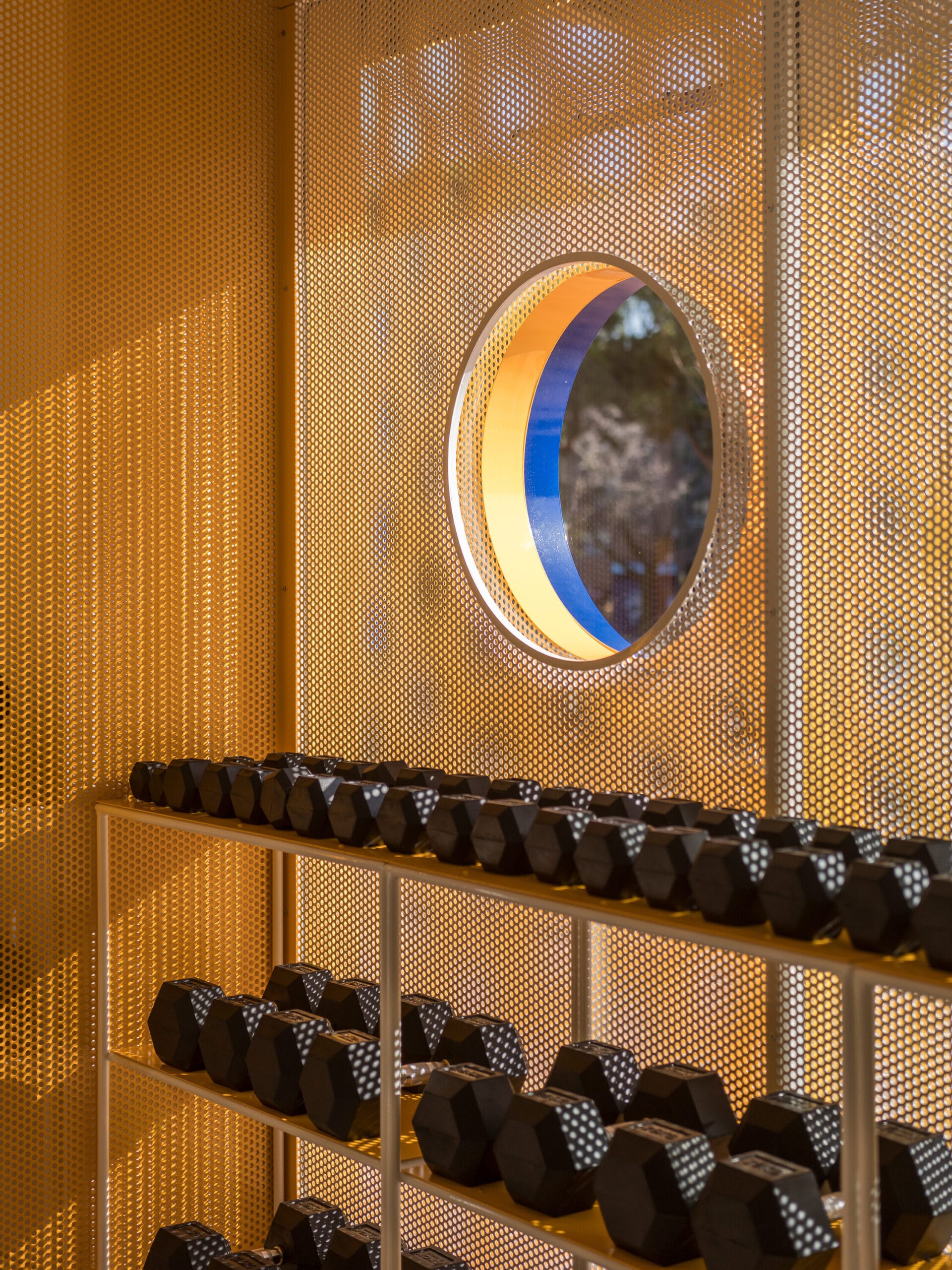 Een rek met zeshoekige dumbbells voor een geperforeerde gele wand met een rond raam