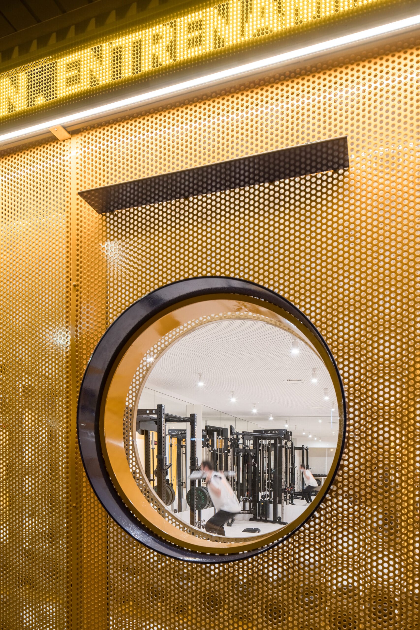 Zicht op een fitnessruimte met krachtstations door een rond raam in een gele geperforeerde wand